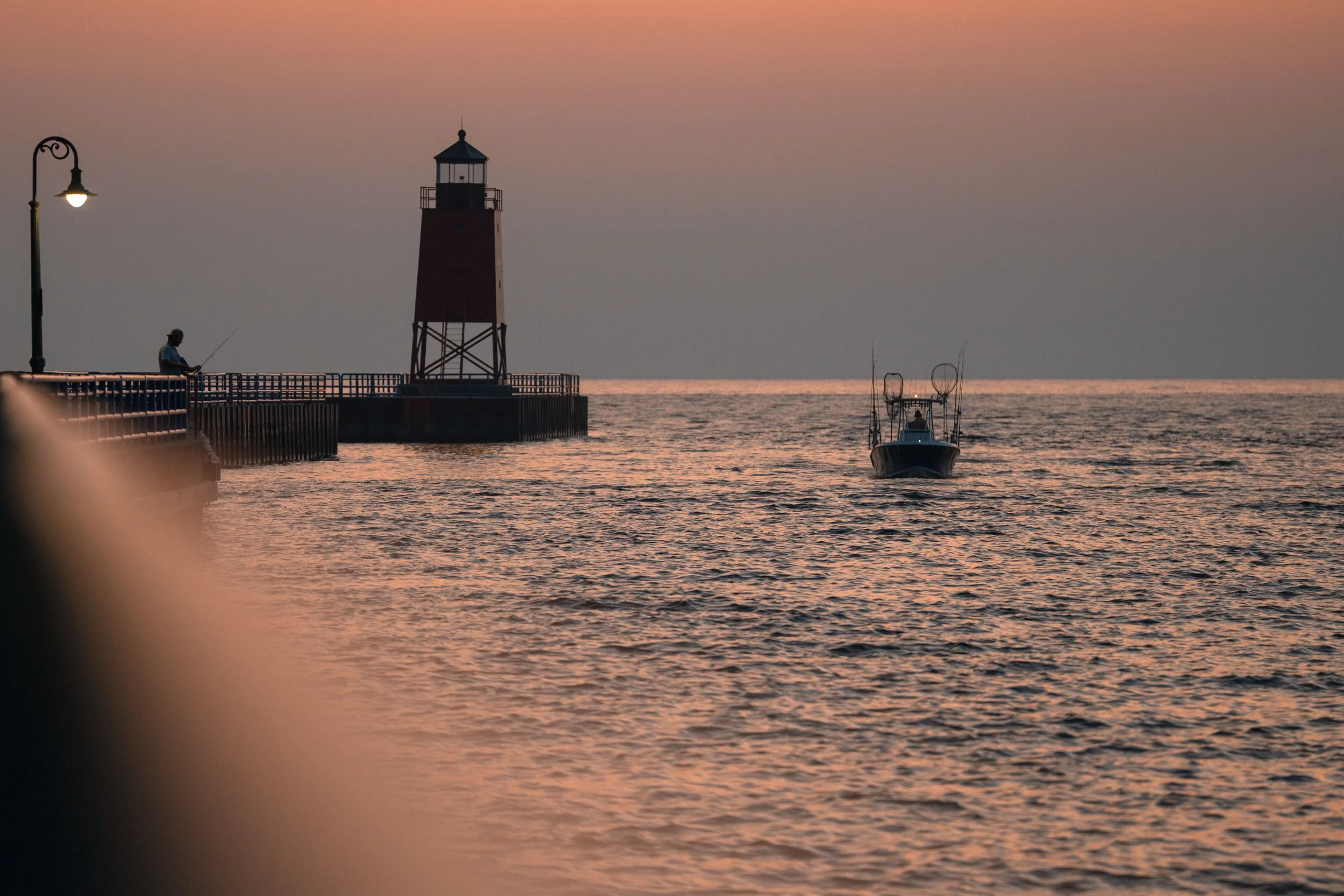 A sunset at the ocean featuring a lighthouse on a pier, a man fishing from the pier, and a boat on the water.