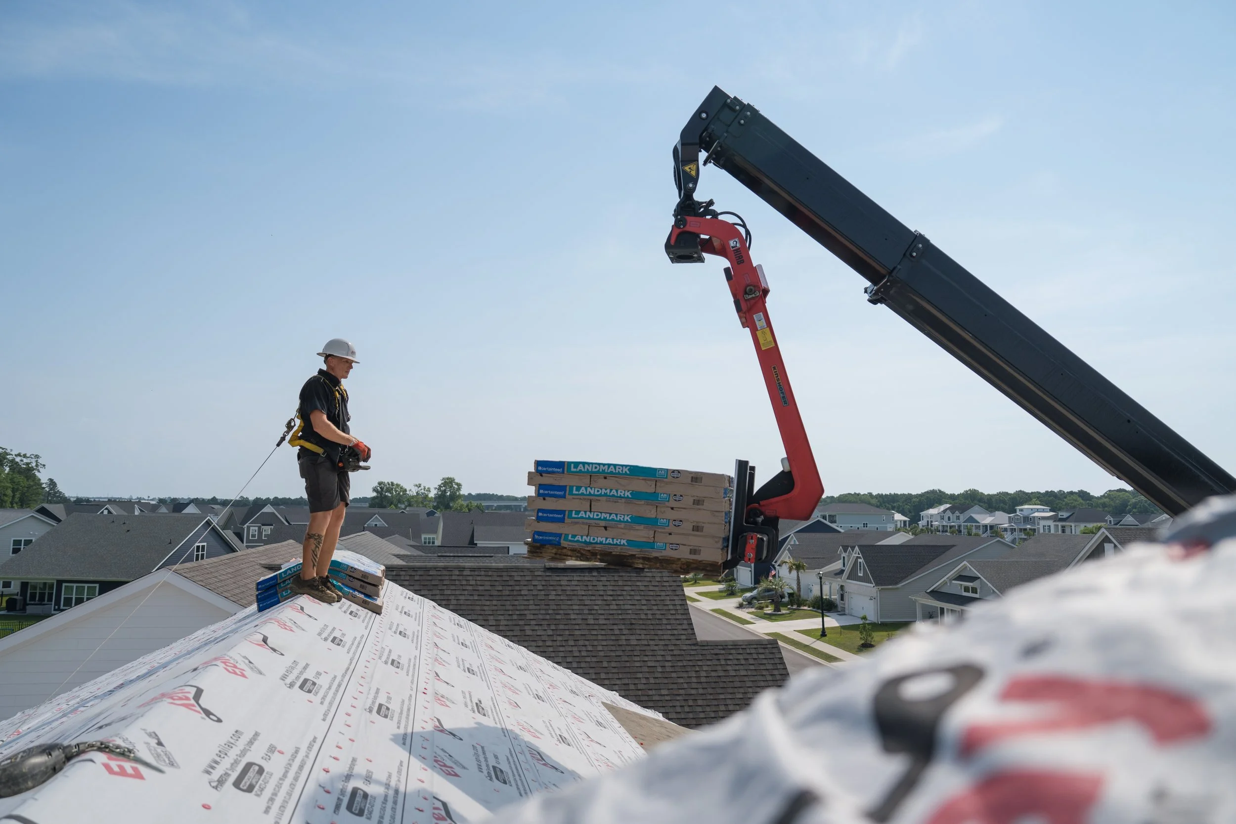 A construction worker standing on a roof installing shingles as a crane lifts packs of shingles to him.