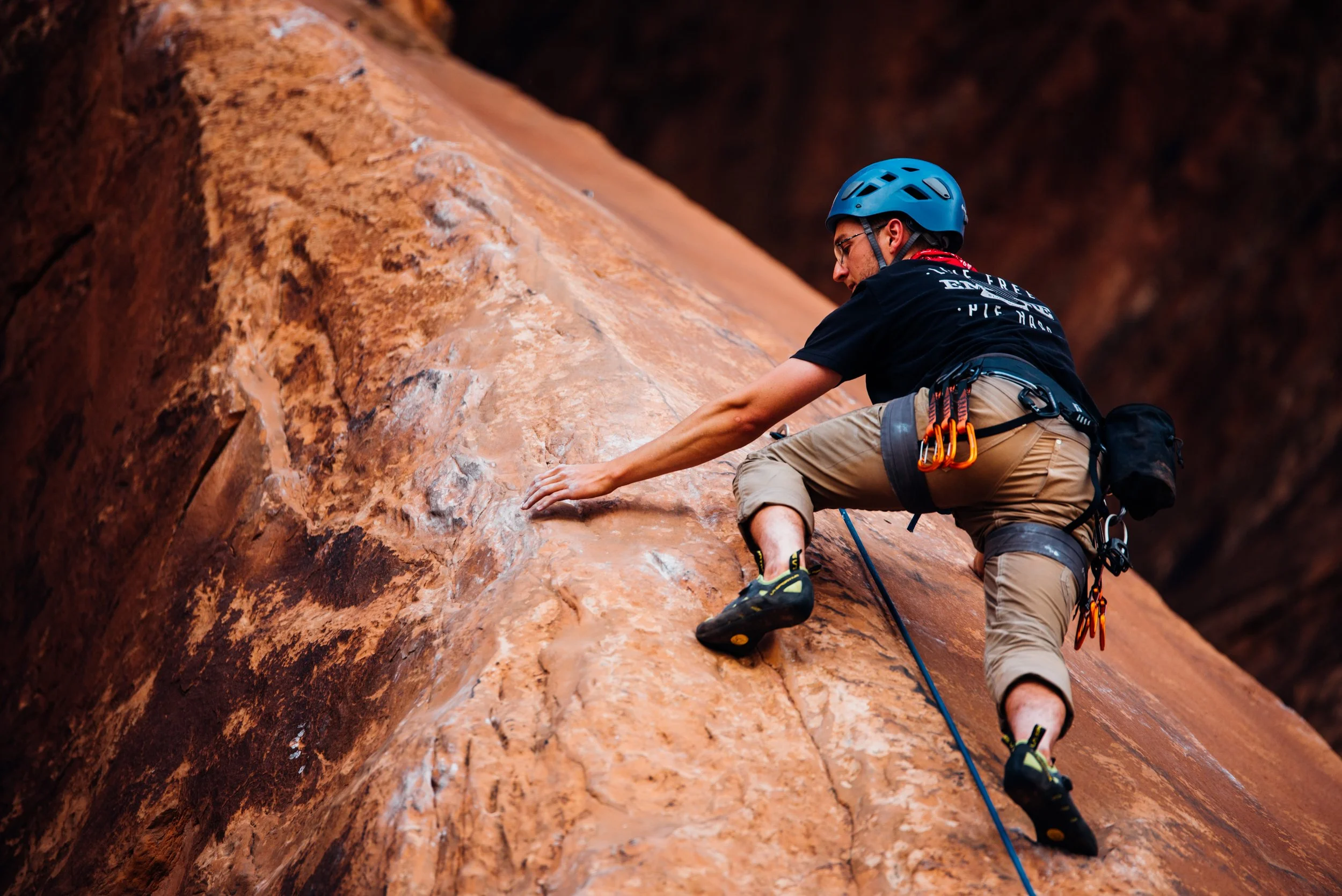 A male rock climber wearing a blue helmet, black t-shirt, and khaki shorts ascending a steep, reddish-brown rock face. He is using climbing gear including quickdraws and a harness, with a safety rope attached for protection.