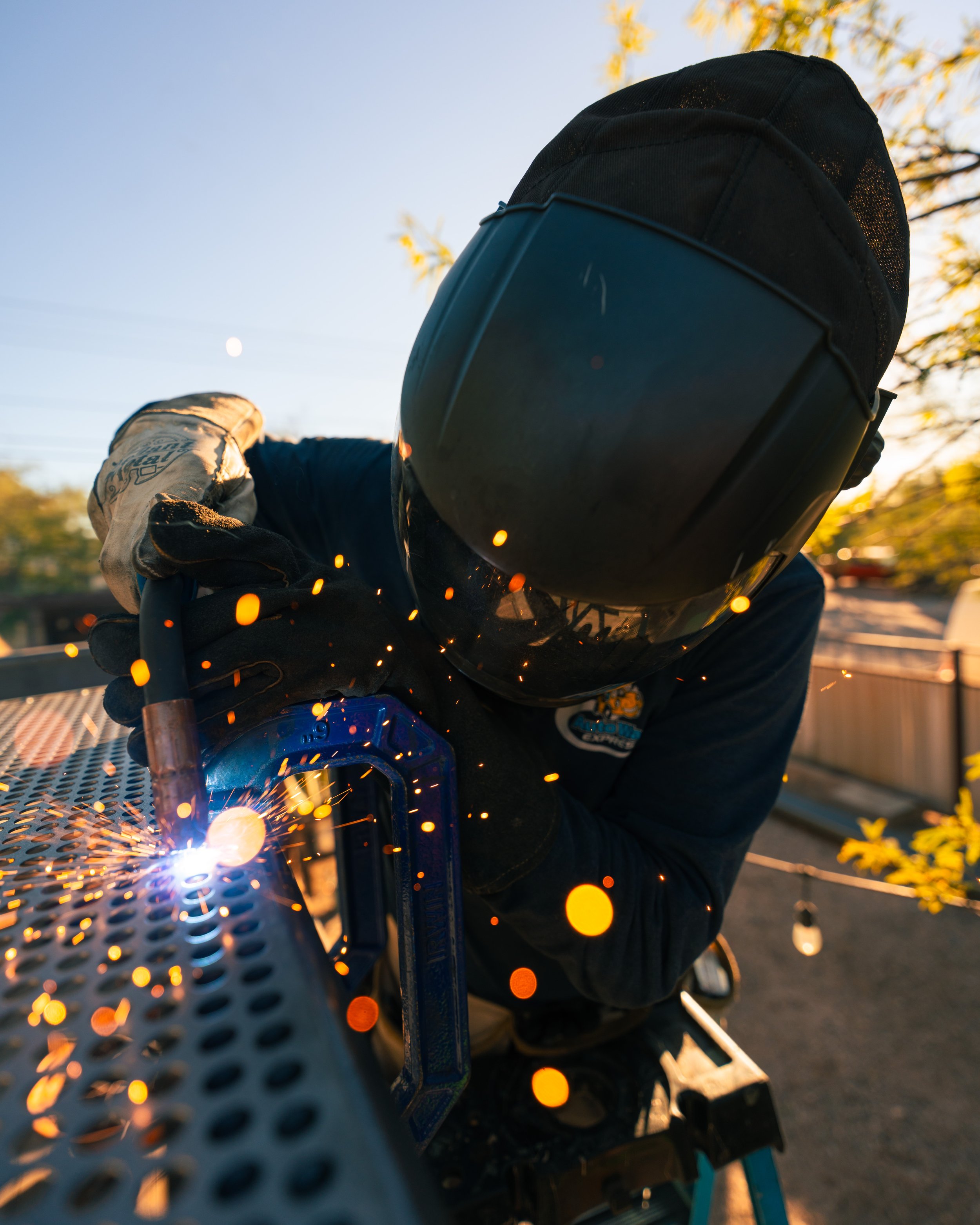 A person welding outdoors at sunset, wearing a protective helmet and gloves, sparks flying from the welding process.