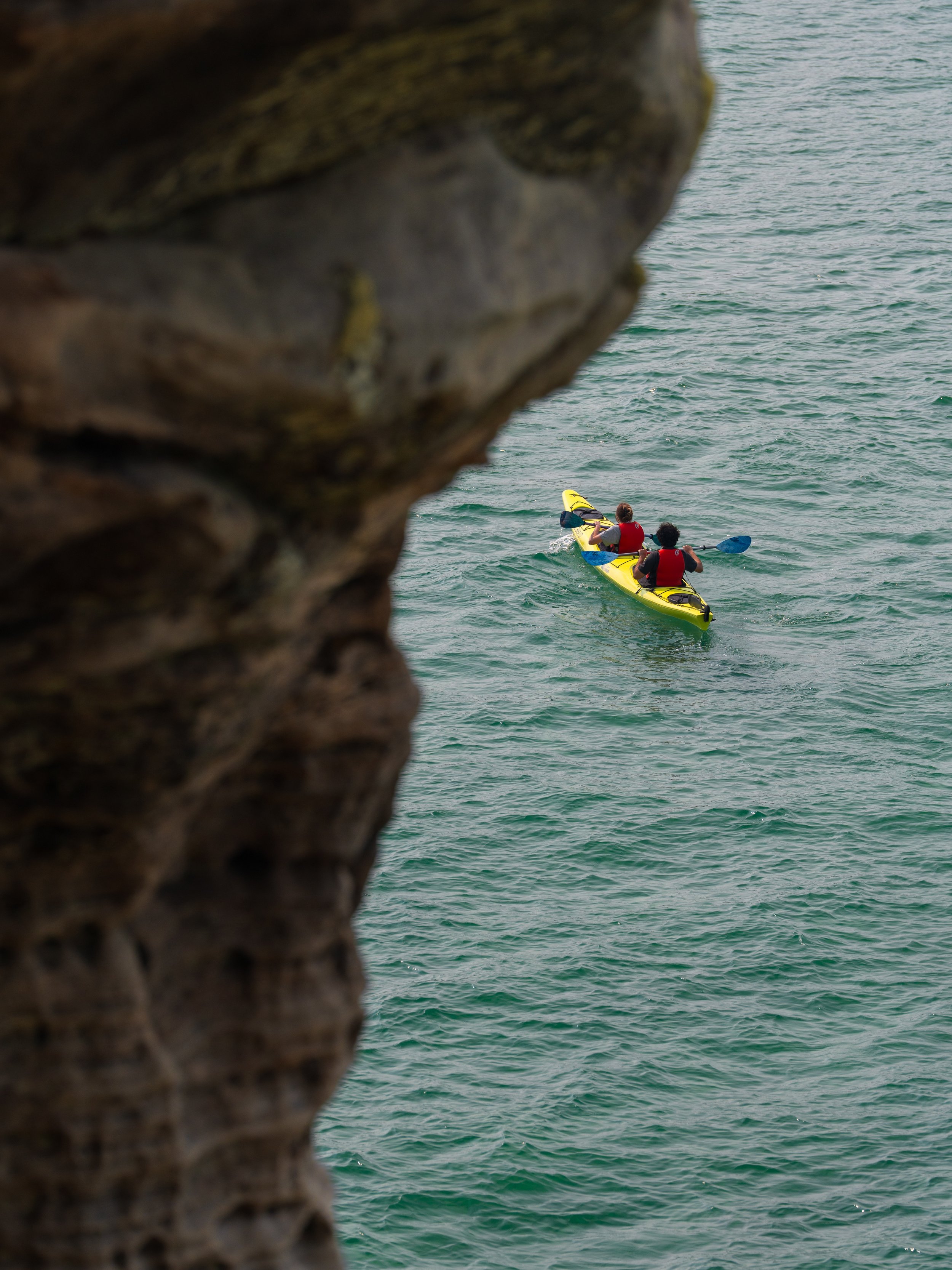 Two people in a yellow kayak paddling in the water, framed by a large rock in the foreground.
