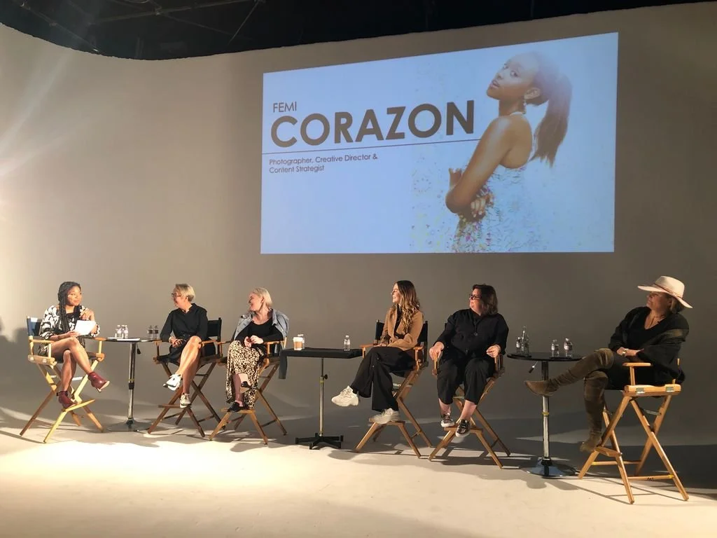 APA Women In Photography - The Female Lens - A panel discussion with six women sitting on stage in director chairs, with a large screen behind them displaying a presentation slide that reads "FEMI CORAZON," with a photo of a Black woman on screen