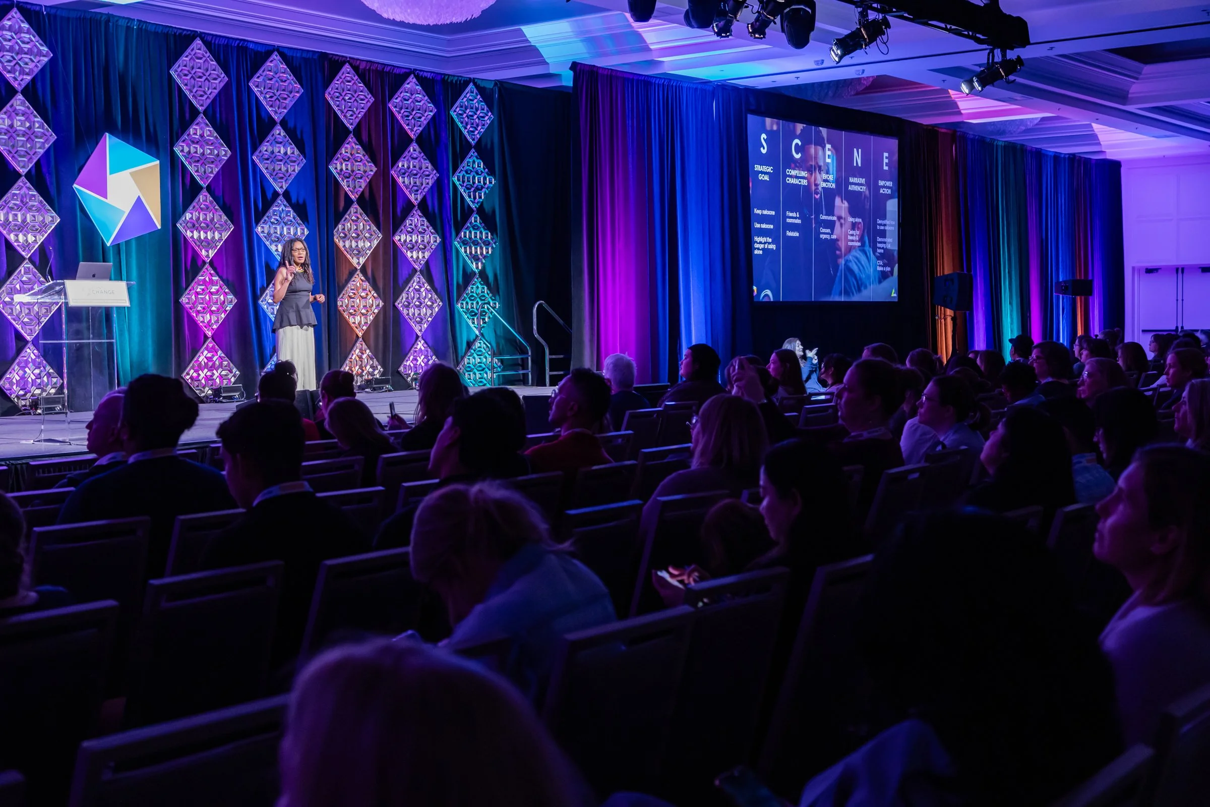 Femi Corazon stands on stage giving a presentation at a conference with a large audience seated in rows. The stage is decorated with colorful geometric shapes and lighting. A screen behind her displays a slide with the word 'SCENE' and various text.