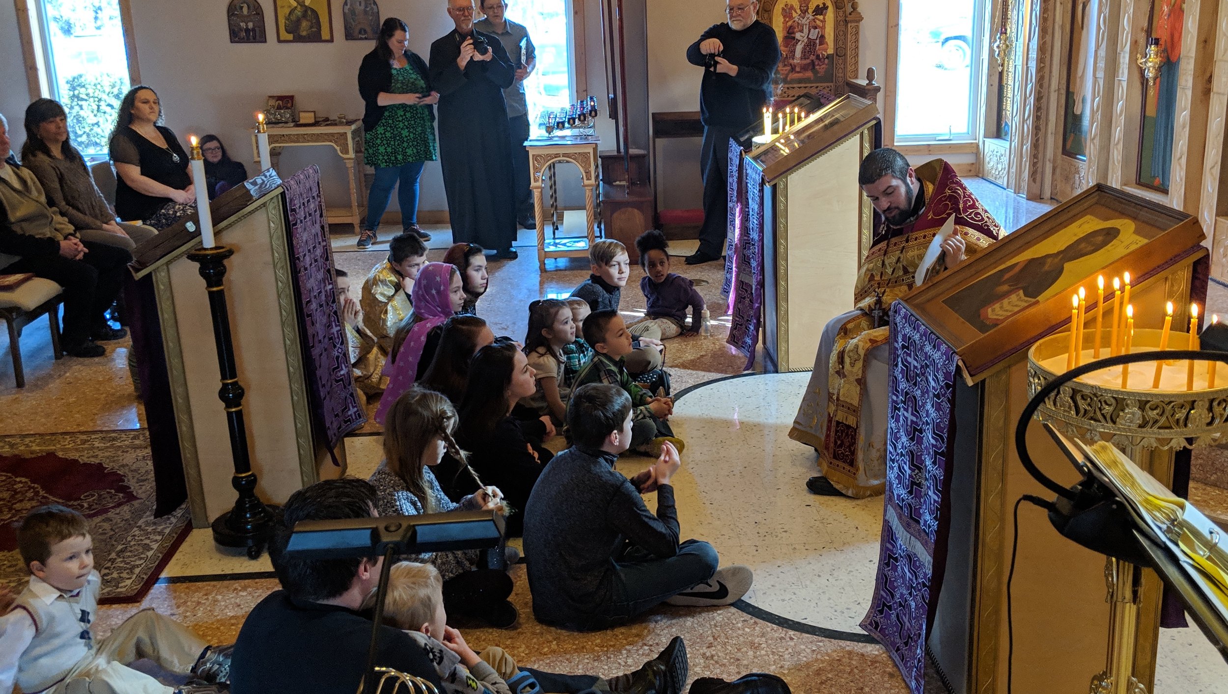  children sitting in front of priest while he reads to them with parents sitting in background 