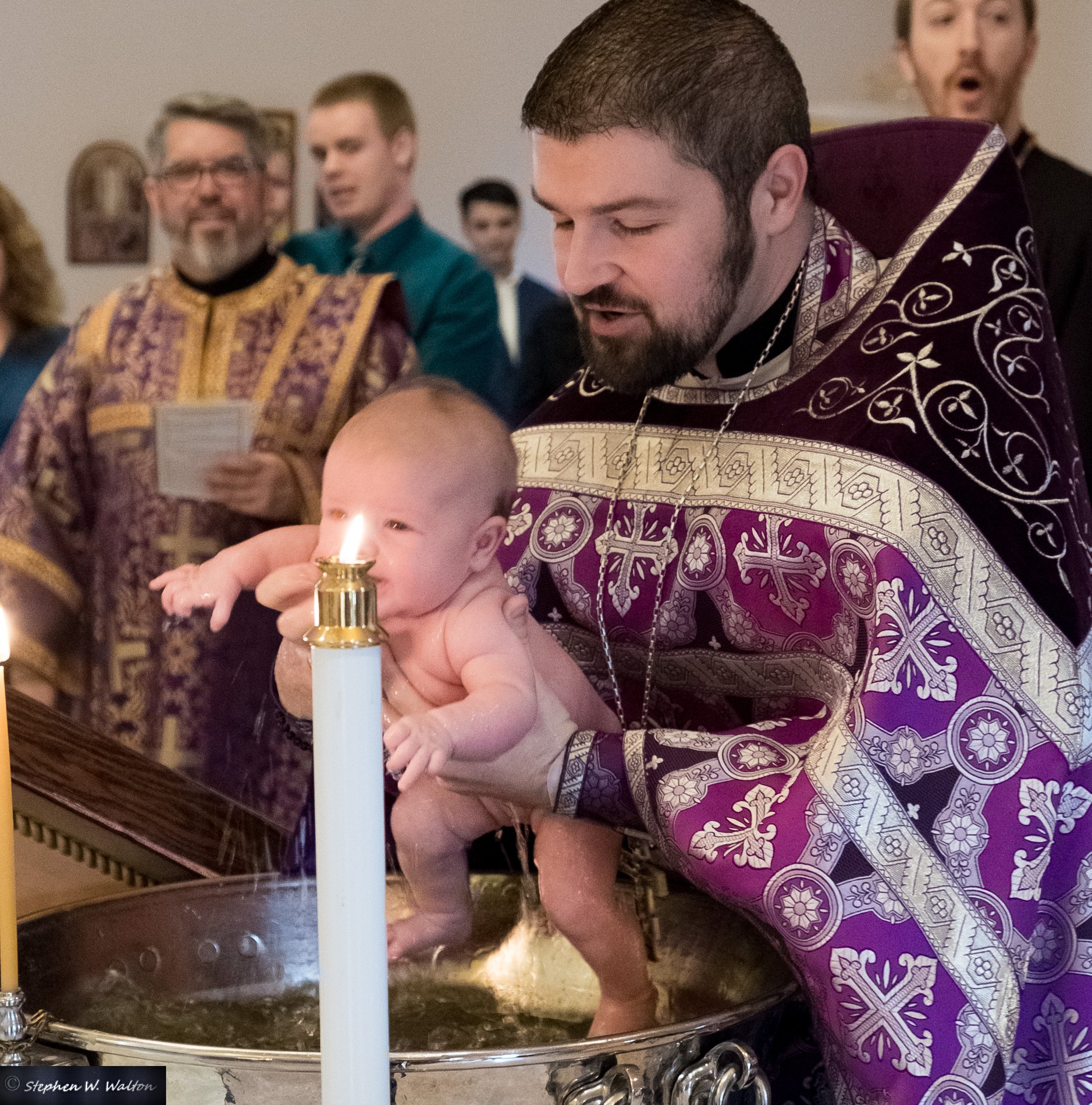 priest baptizing a infant with people in background smiling