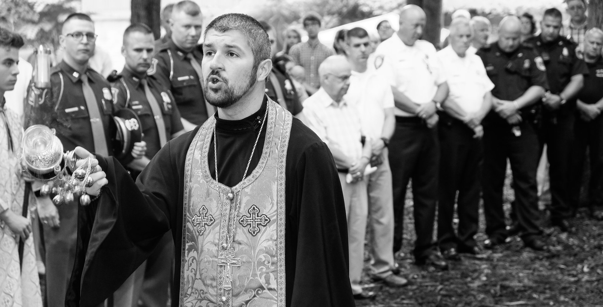 priest censing and praying with first responders in background black and white 