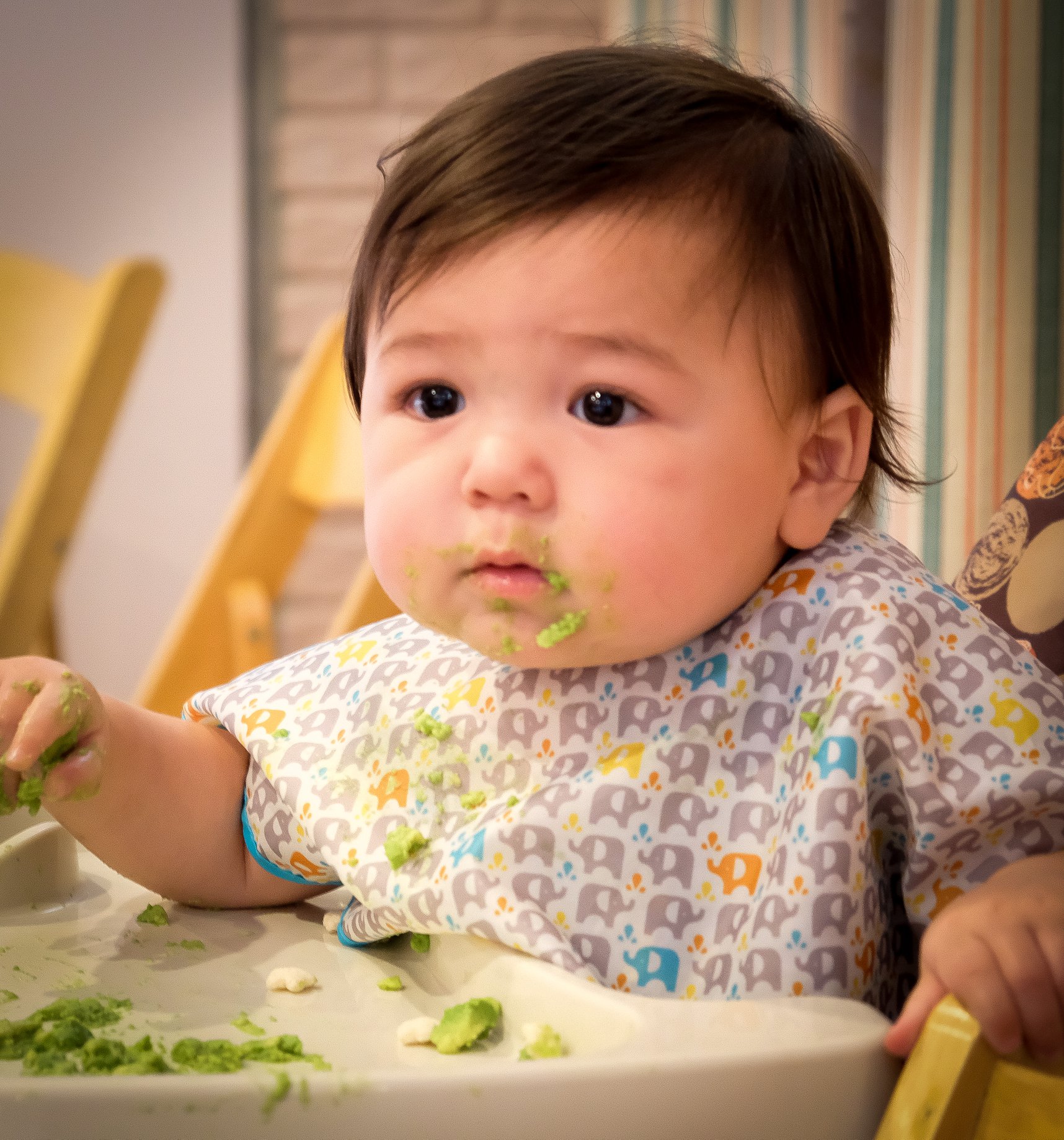  baby girl  in high chair eating with green peas on chin 