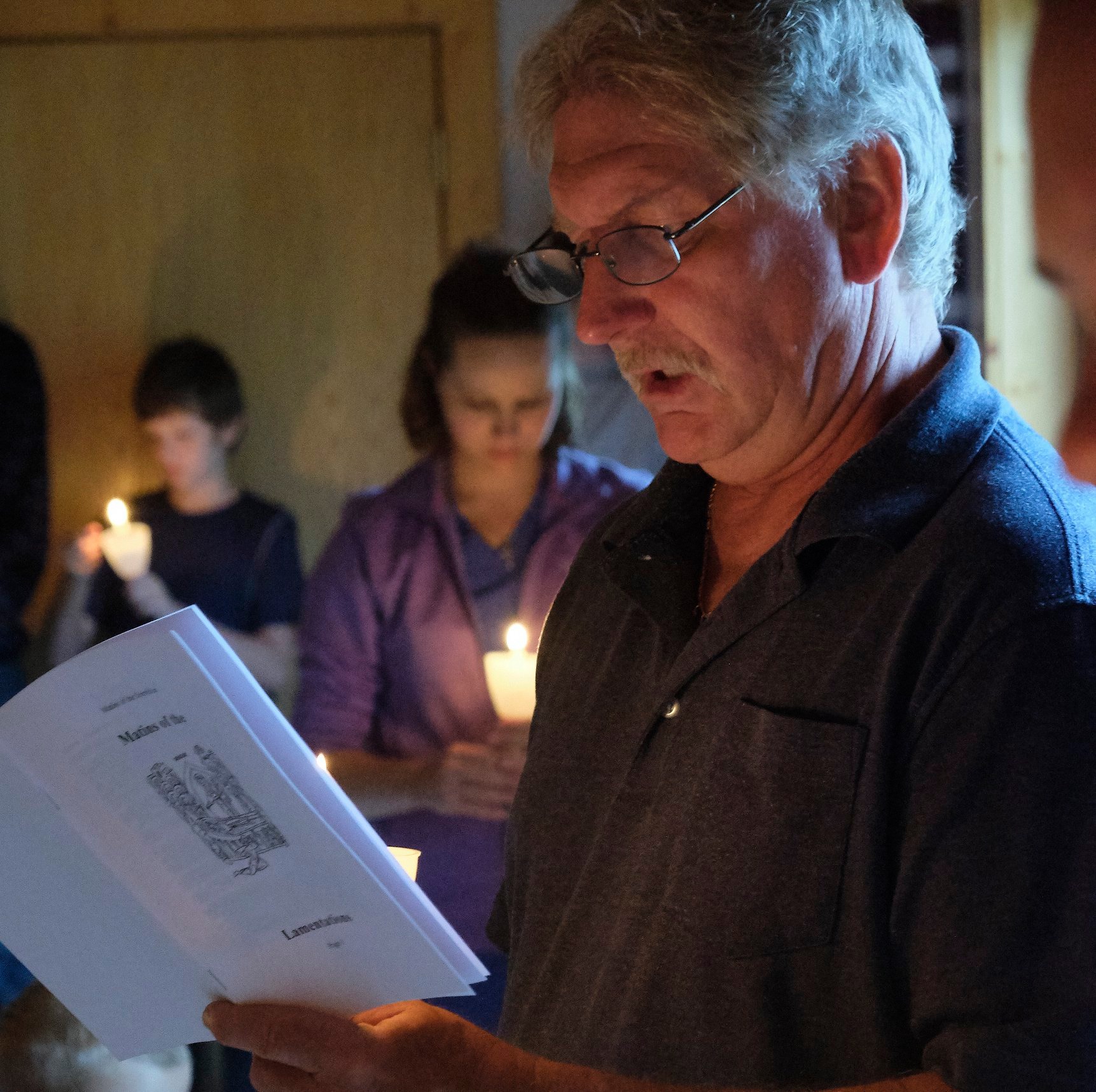  Man with glasses and grey hair reading from a service book with woman and young boy in background holding candles. 