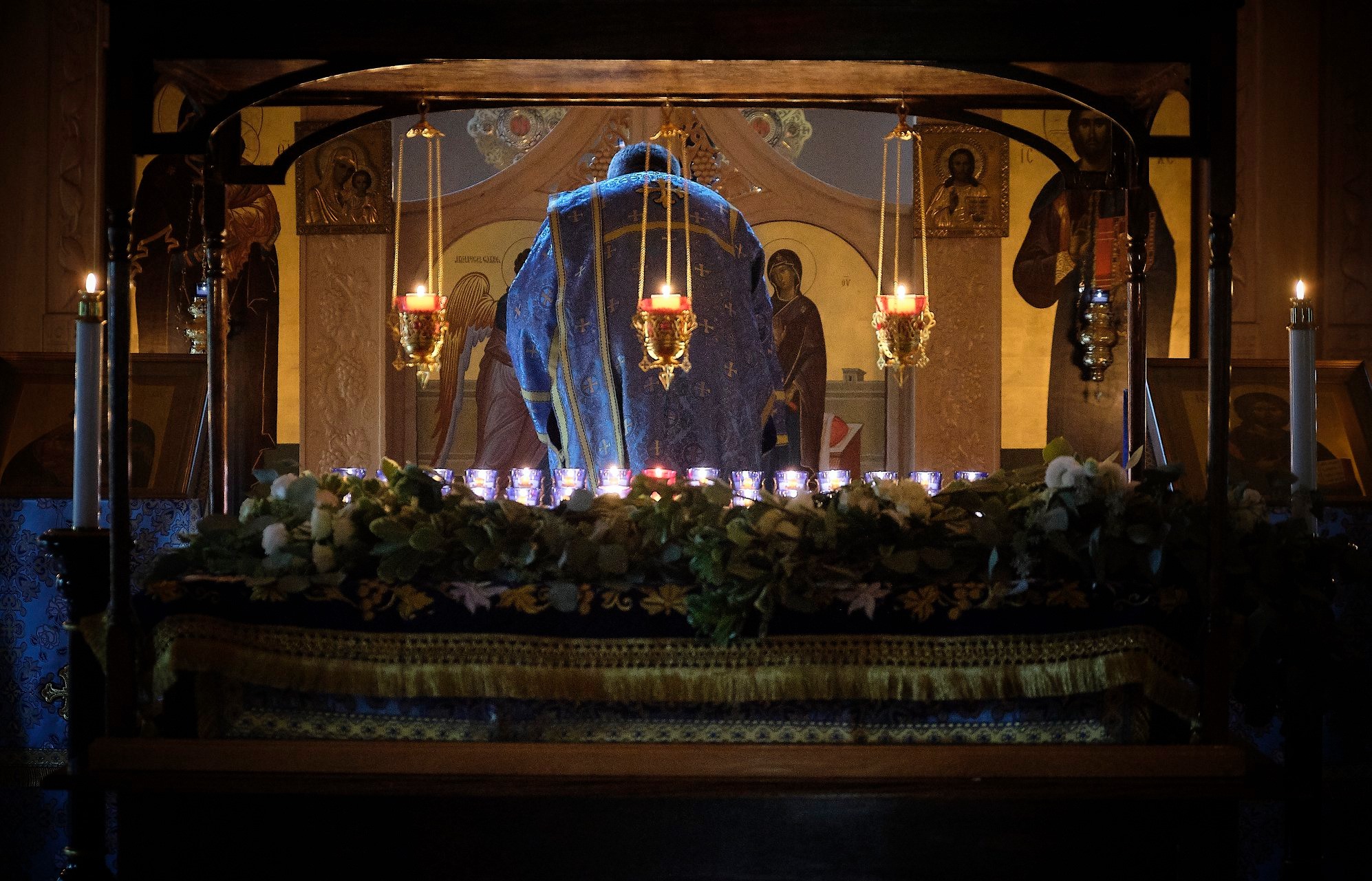  Looking through the ceremonial tomb that has lit candles and flowers inside. Deacon is shown from behind bowing in front of the altar.  