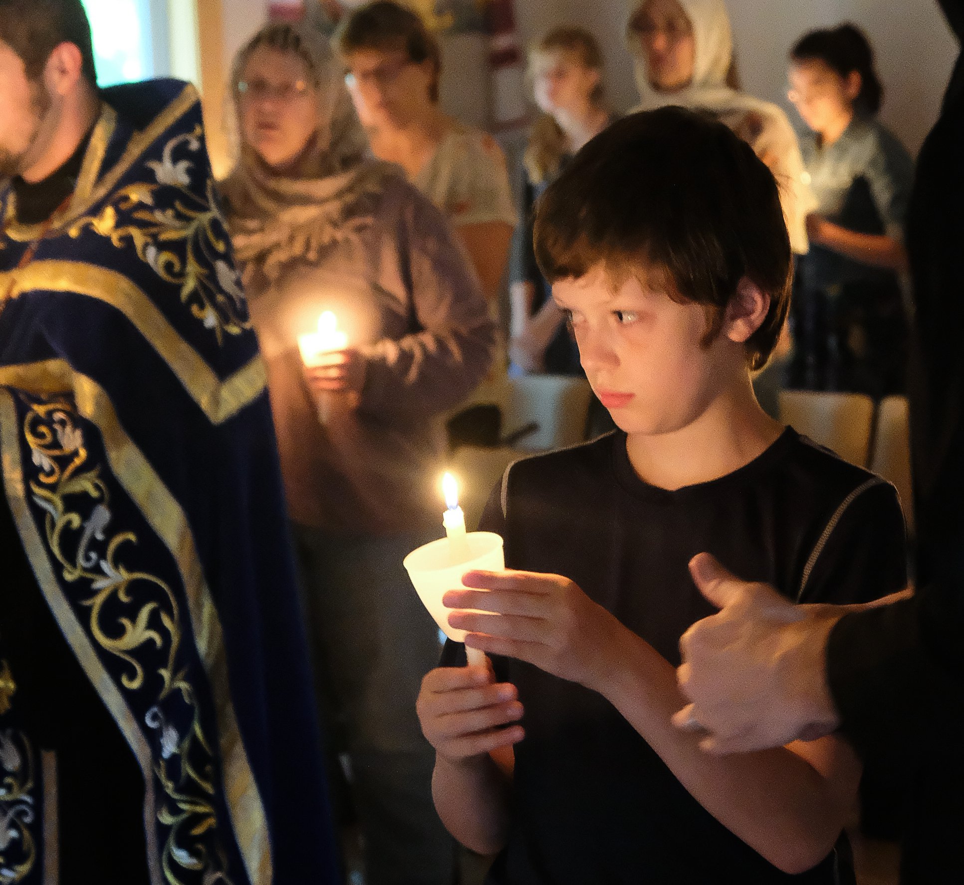  Young boy looking forward while holding a candle. Priest and other parishioners are in the background all facing the same way.  