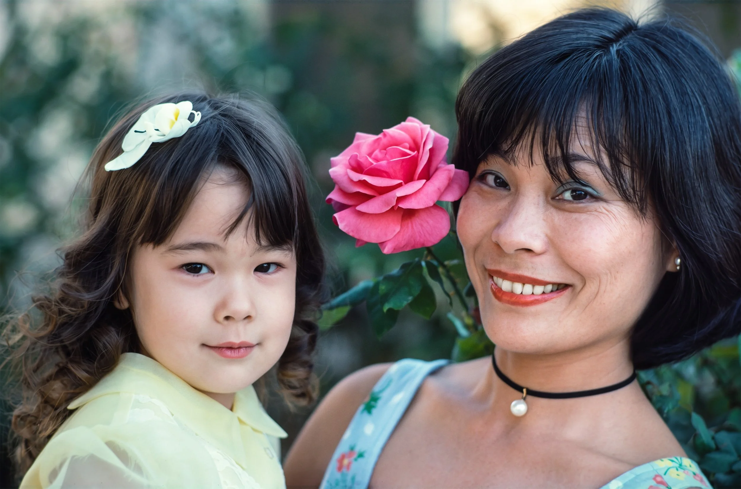 Mother and four year old daughter pose for a natural light photograph near a rose bush offering a timeless moment captured.