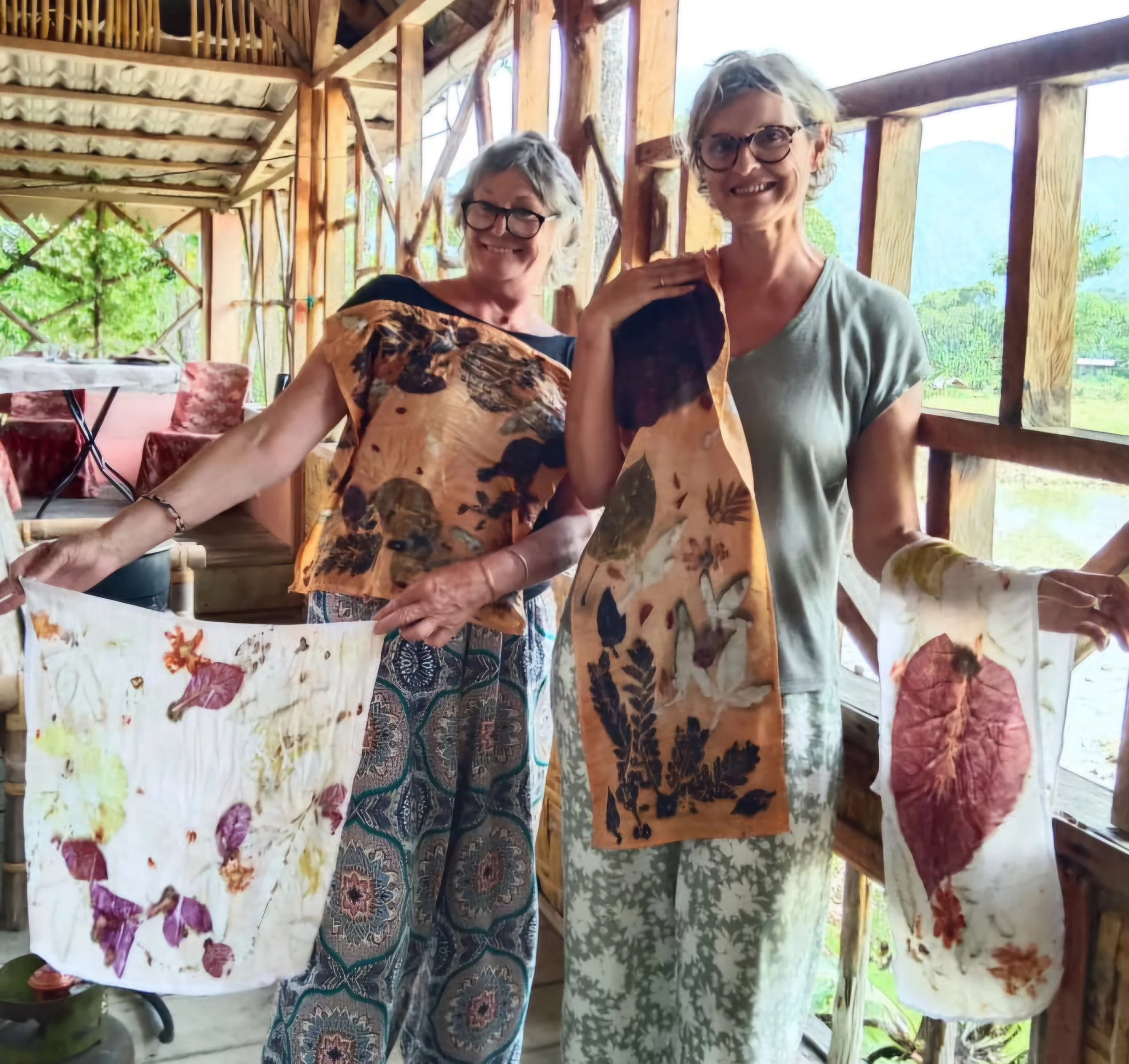 Two women display handmade fabric banners with pressed leaves and flowers inside a wooden structure with a scenic outdoor view.