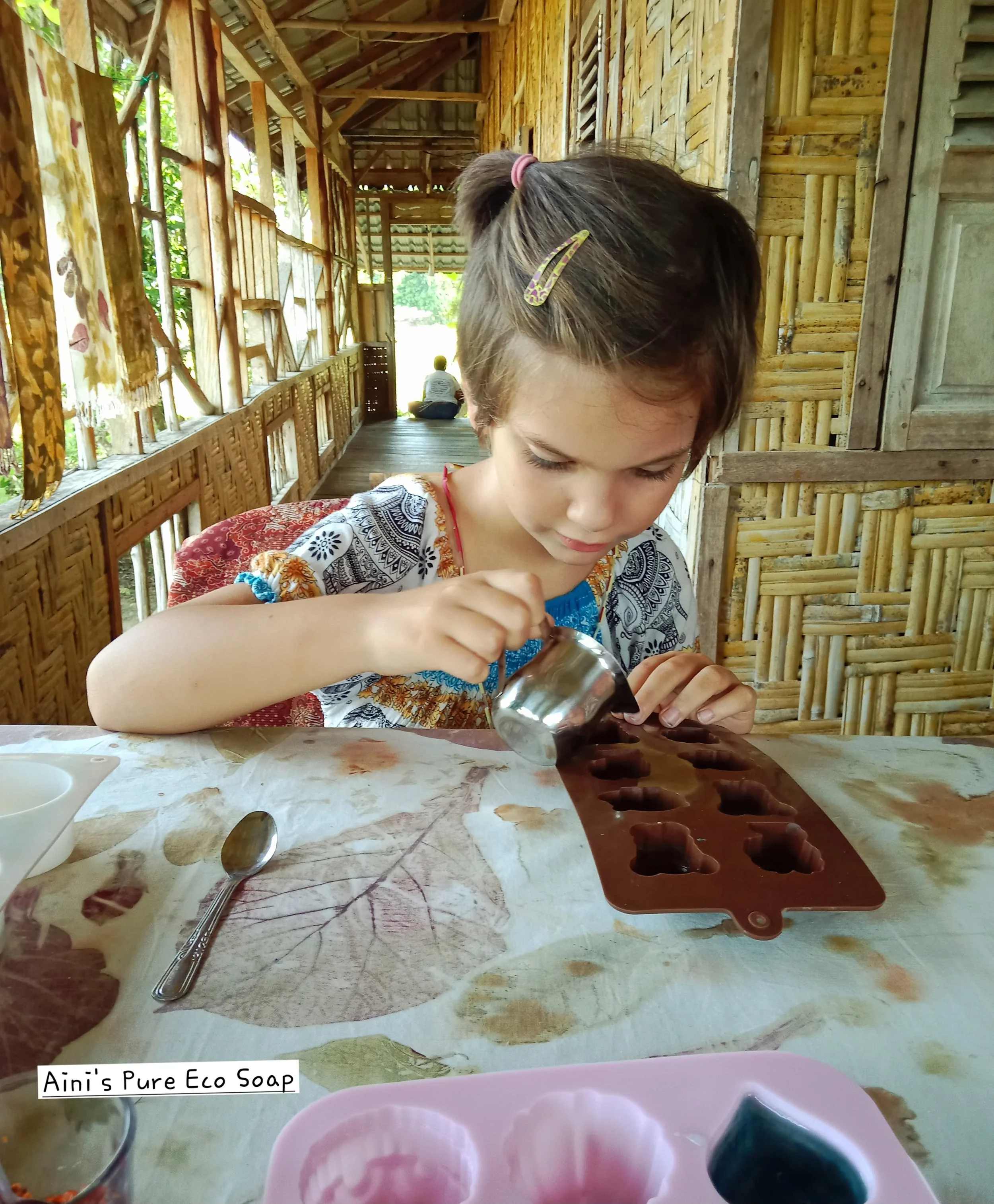 A young girl with a hair clip and colorful shirt is pouring a liquid into a silicone mold for making soap on a table with leaves printed on a tablecloth.