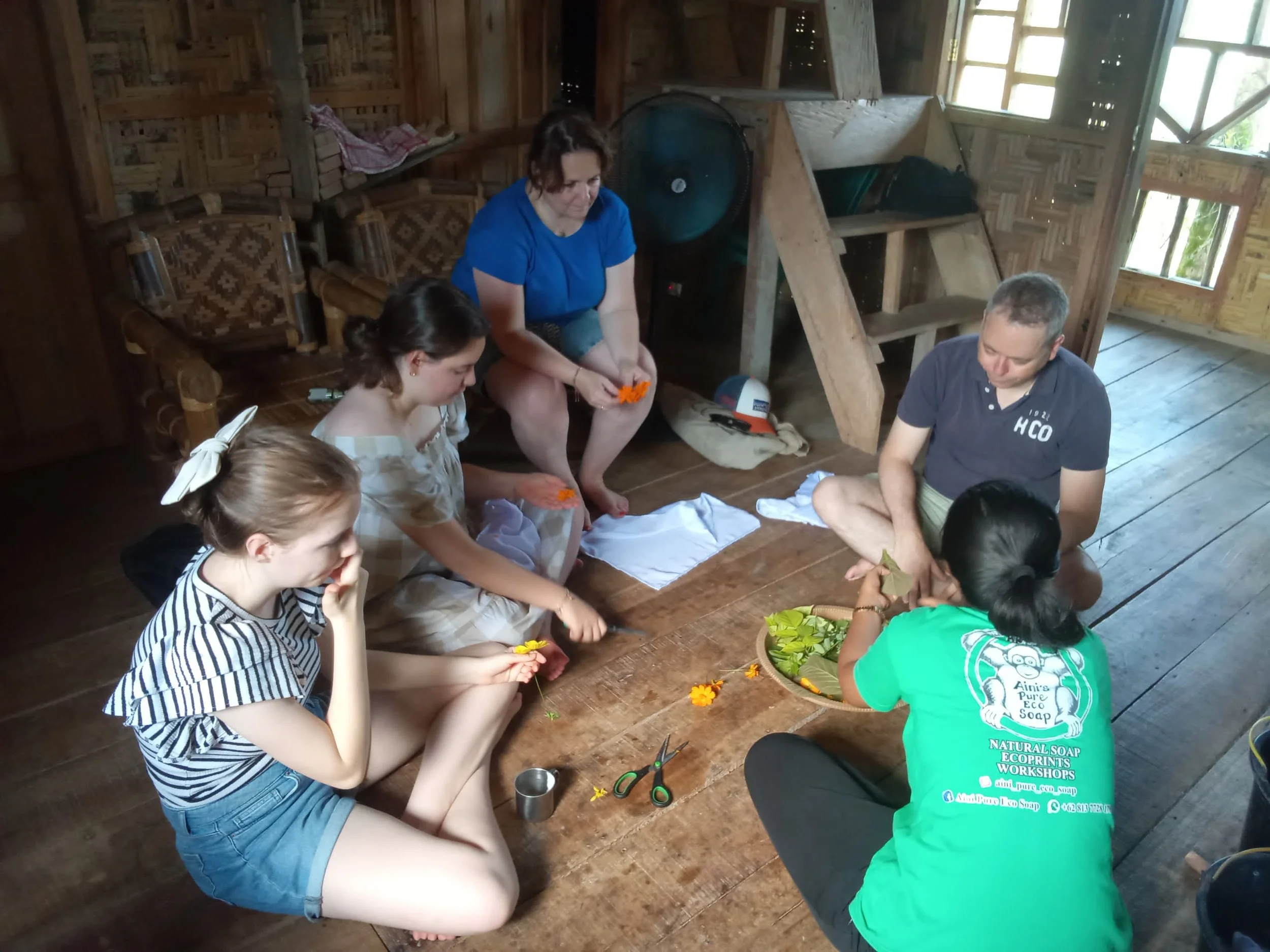 A group of six people sitting on the wooden floor of a rustic room, engaged in a craft activity involving orange flowers. There are scissors and other craft materials on the floor, and they appear focused on their task. The room has wooden walls, a window allowing natural light, and some shelves and chairs in the background.