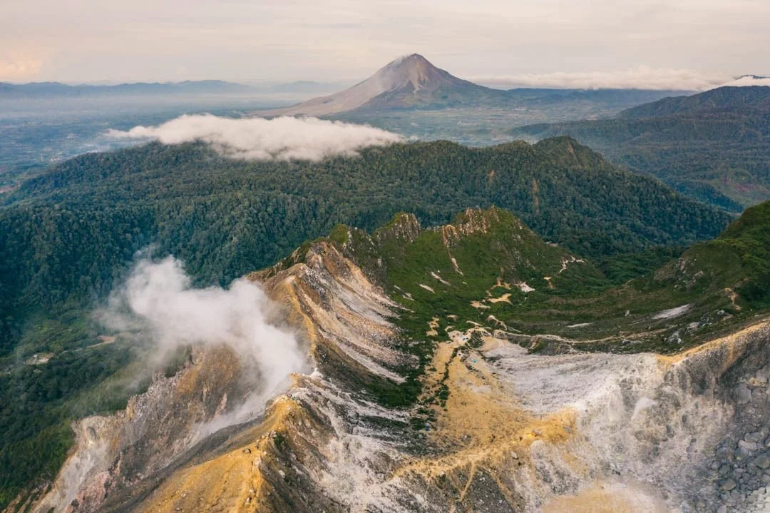 Aerial view of a volcanic landscape with a mountain in the background, lush green hills, and steam rising from the volcanic crater in the foreground.