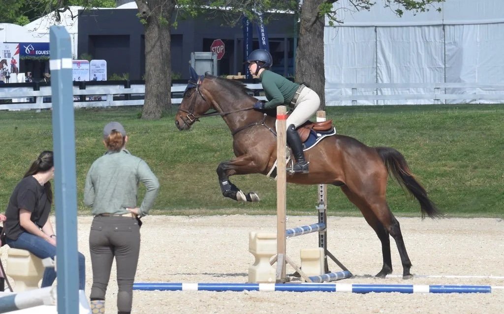 A rider on a brown horse jumping over a blue and white obstacle in a riding arena, with two women observing nearby.