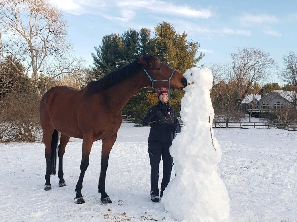 A person standing outdoors in snow, wearing a black jacket and red hat, smiling, next to a large snowman. There is a horse with a blue halter leaning in to nuzzle the snowman. Trees and a house are in the background under a partly cloudy sky.