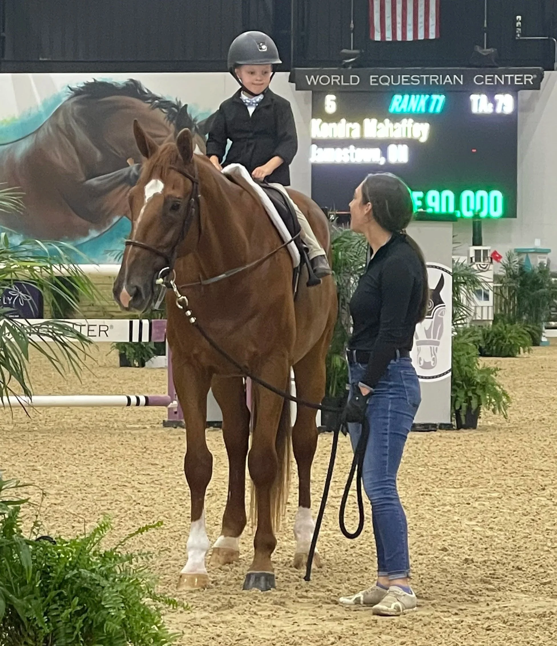 A young boy in riding gear sitting on a brown horse in an indoor riding arena at the World Equestrian Center, with a woman holding the horse's reins and a digital scoreboard in the background.