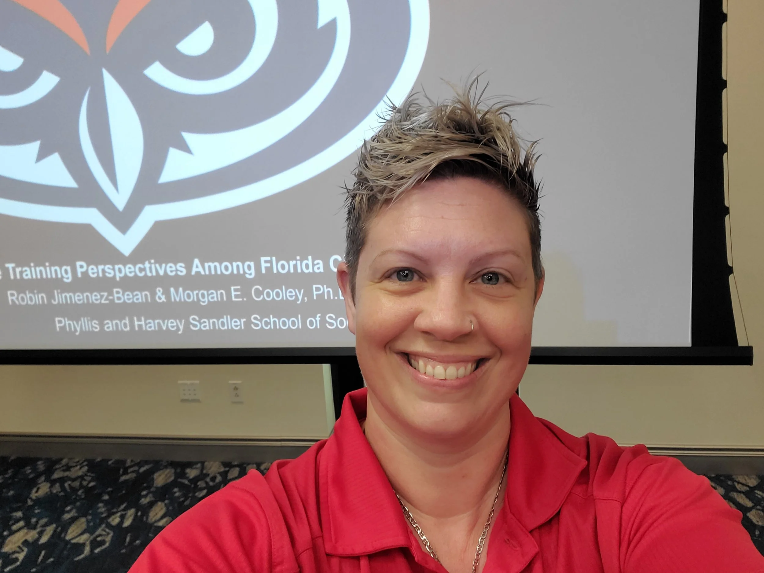 A woman with short, spiky blonde hair taking a selfie in front of a presentation slide. She is wearing a red shirt and smiling. The slide behind her displays a large logo with circular and floral design elements and the title 'Training Perspectives Among Florida...'
