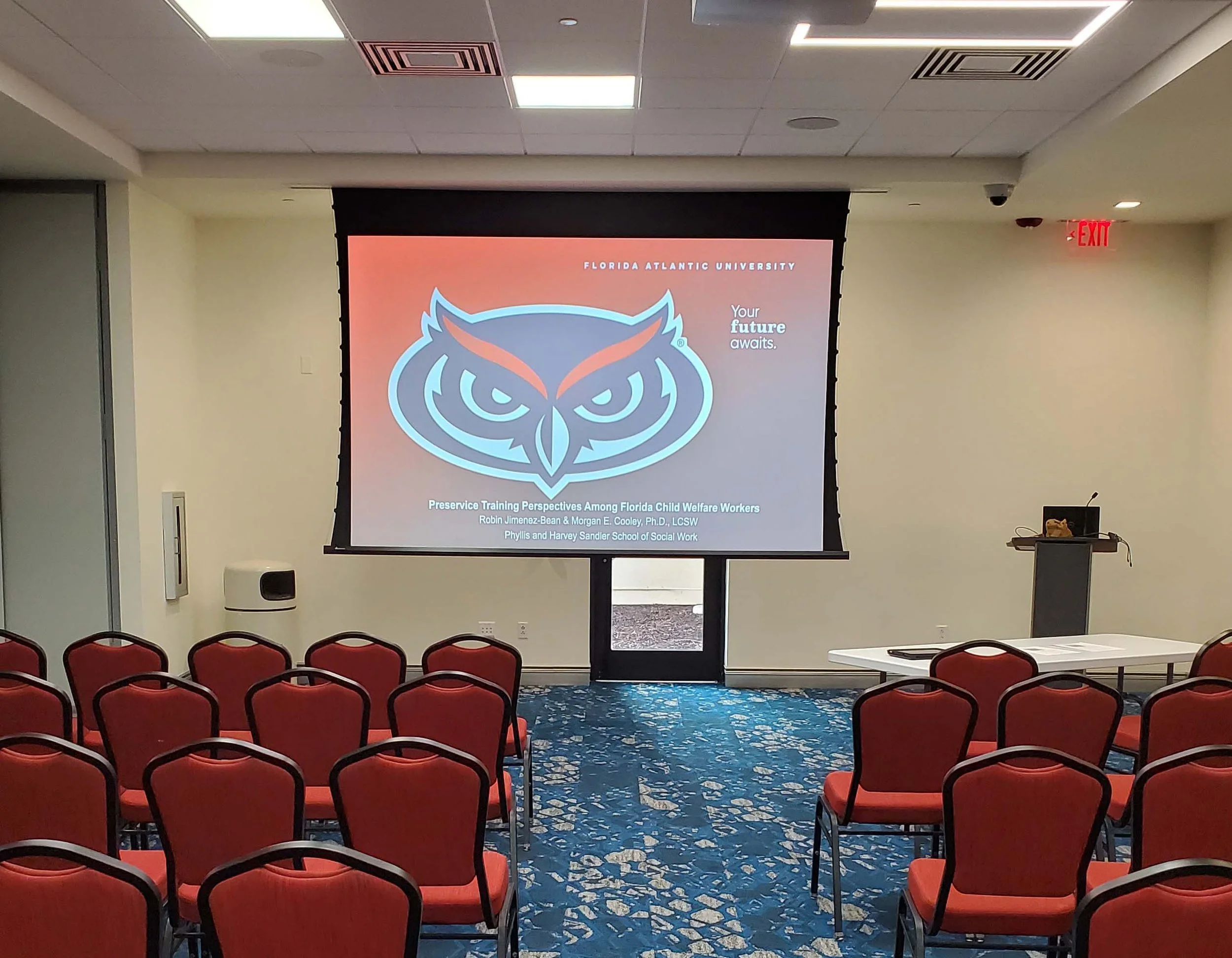 Conference room with red chairs arranged in rows facing a large screen displaying a presentation related to Florida Atlantic University, with the university's owl logo and the title 'Preservice Training Perspectives Among Florida Child Welfare Workers'.