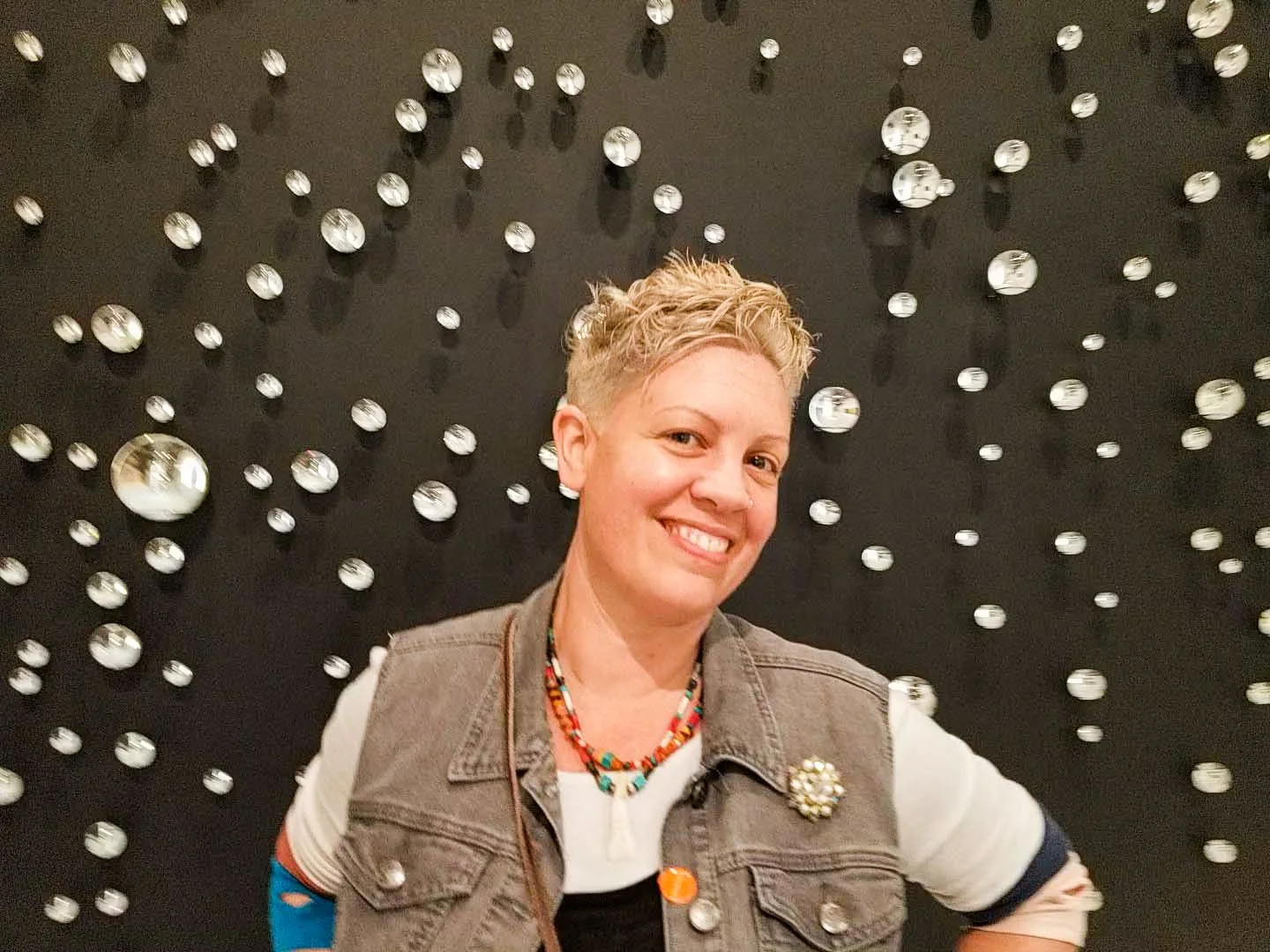 A woman with short blonde hair smiling at the camera, wearing a multicolored necklace, a white t-shirt, a gray vest with a decorative brooch, in front of a black wall decorated with numerous small, reflective, metallic spheres.