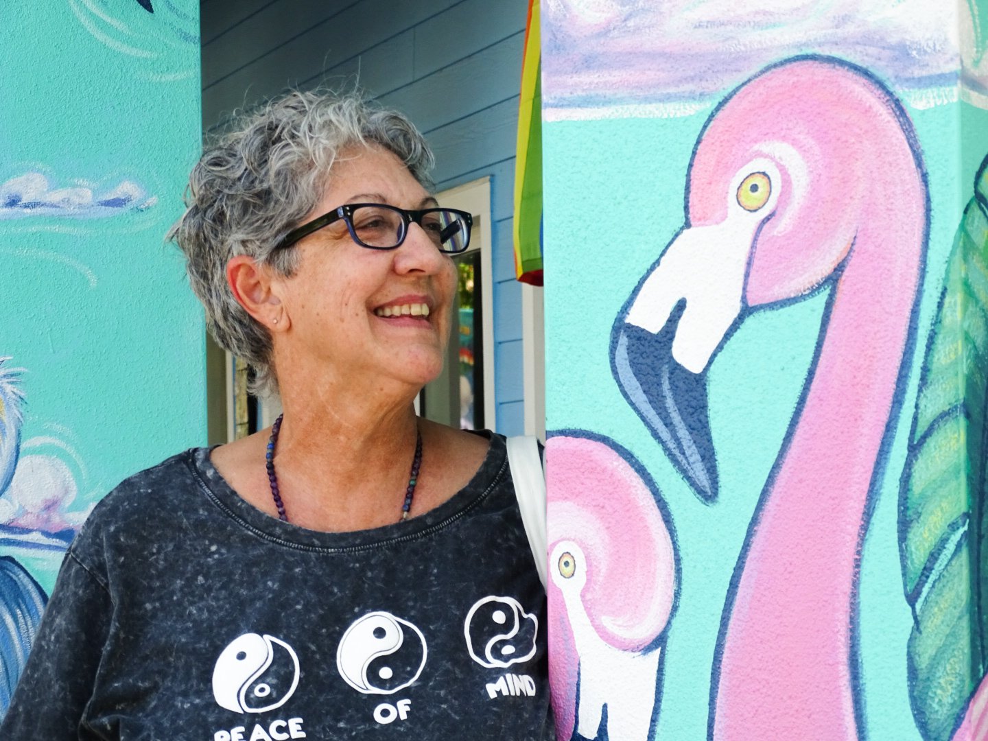 A smiling woman with glasses and short curly gray hair standing next to a colorful mural of a pink flamingo and a baby flamingo.