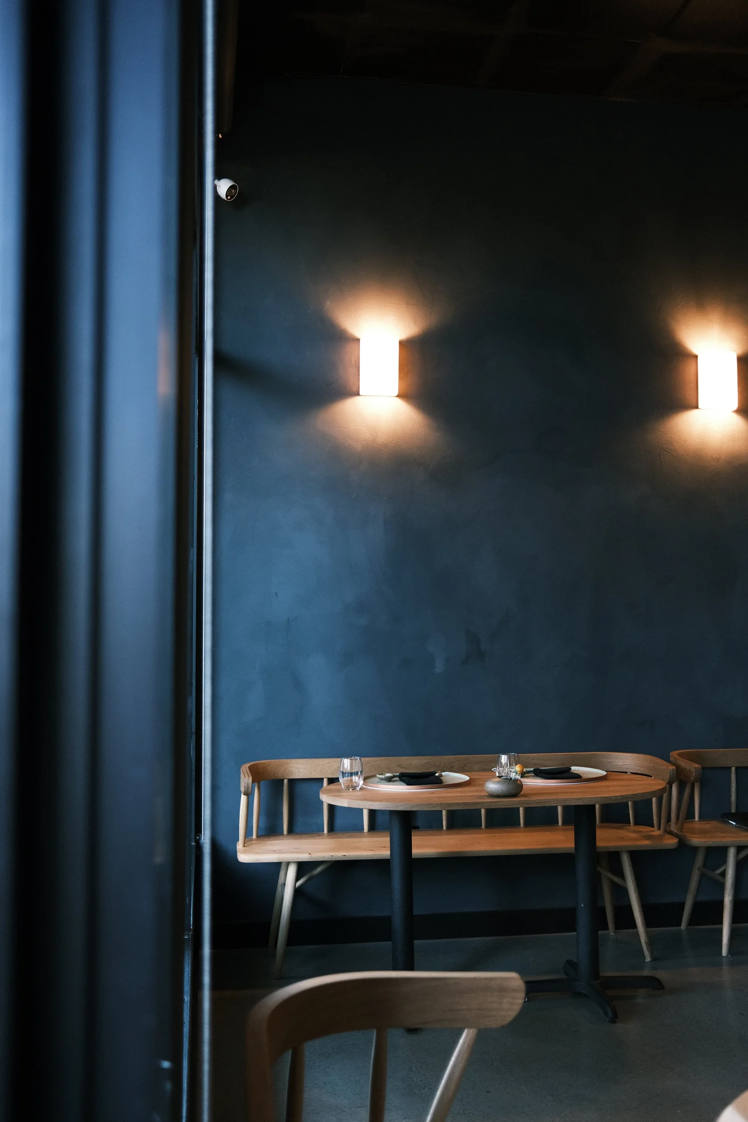 White oak bar seating beneath brass sconces against a deep blue plaster wall at Okesutora, Sacramento