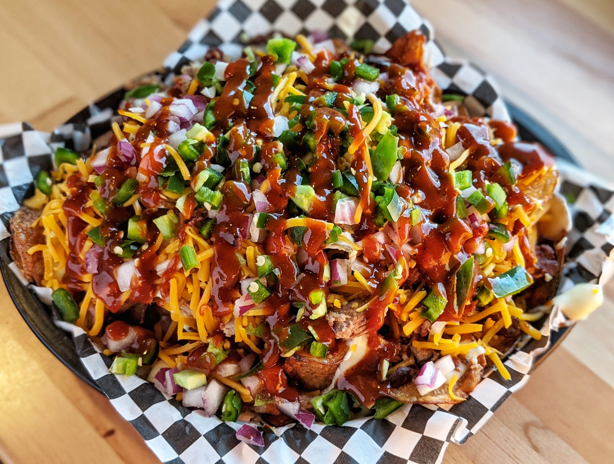 Loaded potato tot bowl topped with cheese, chopped green onions, diced onions, barbecue sauce, and other toppings on a checkered paper-lined tray.