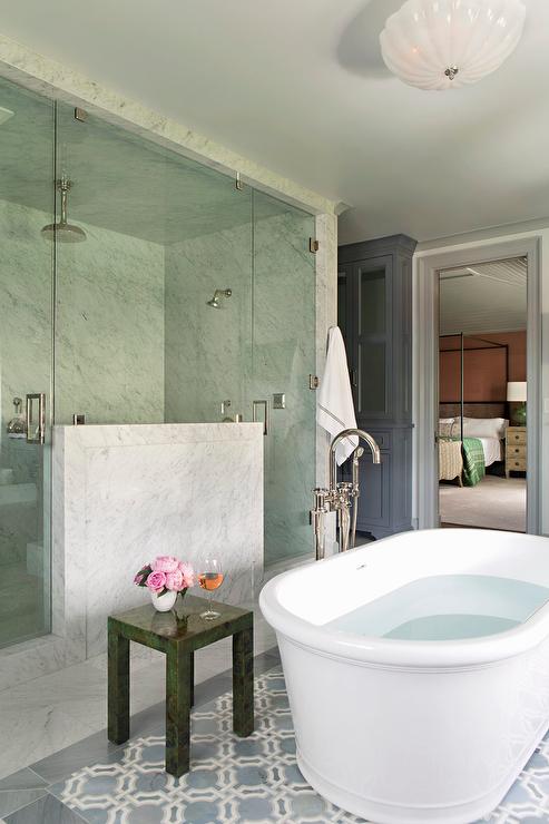 An oval freestanding bathtub paired with a floor-mount tub filler as well as a malachite accent table sits atop a blue geometric tiled floor. A freestanding bathtub is placed in front of a seamless glass and marble walk-in steam shower fitted with his and hers shower heads.
