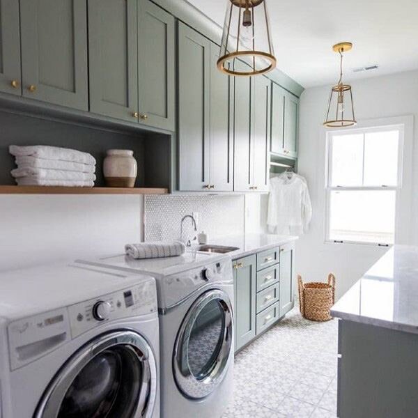 Green gray laundry room is lit by a brass and glass lantern hung in front of green gray shaker cabinets by  MAWR Architecture