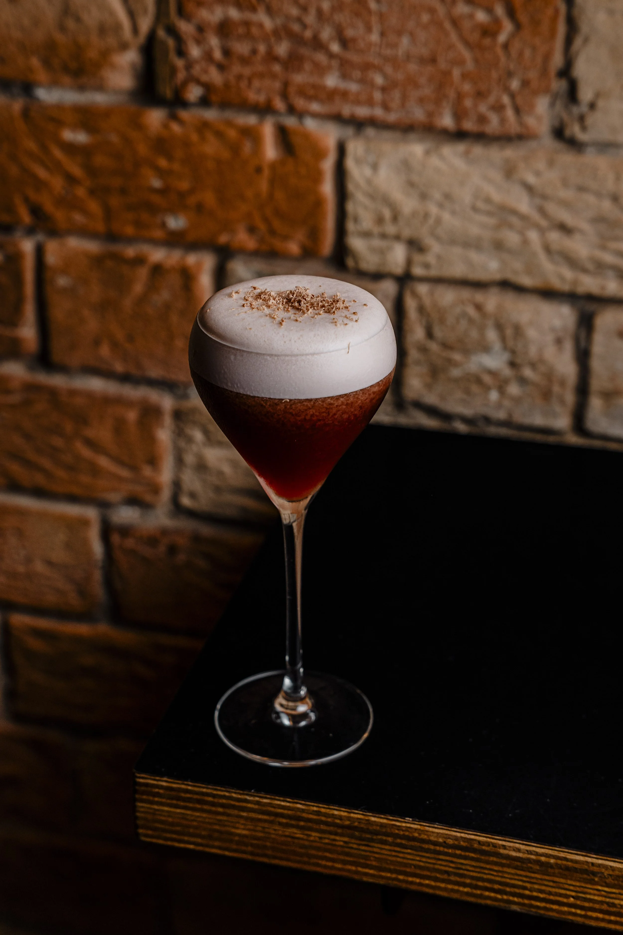 A cocktail in a tall, elegant glass with a layered appearance, topped with whipped cream and sprinkled with cocoa powder, set against a rustic brick wall background.