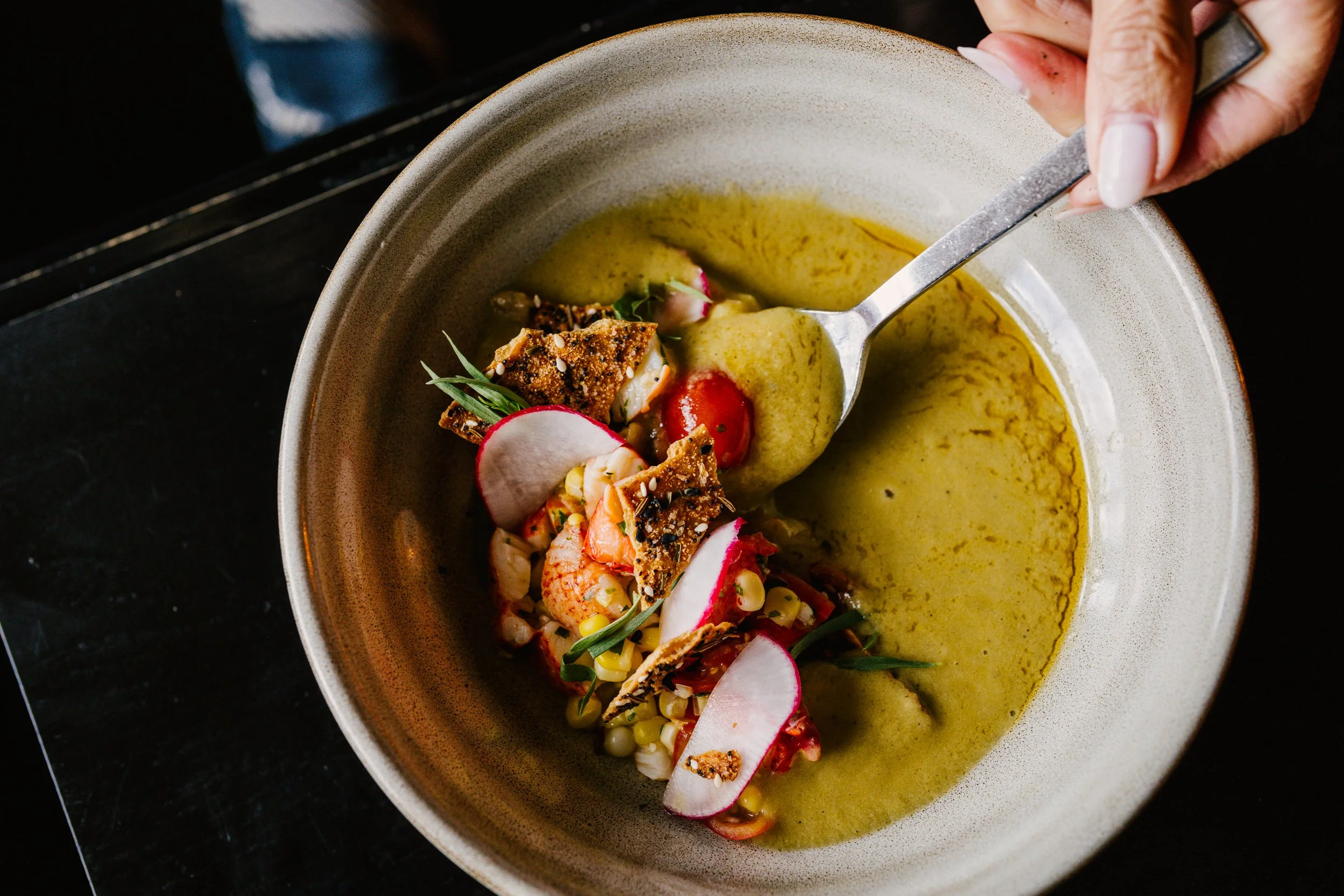 A creamy yellow soup is being spooned up with delicious looking fresh veggies and shrimp