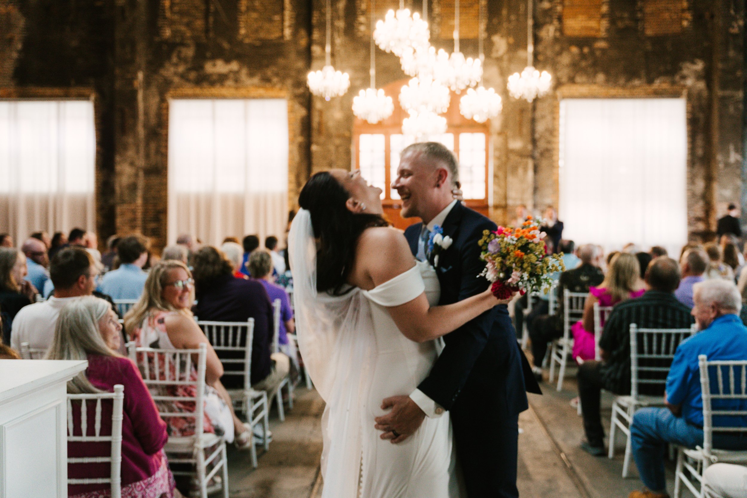 A bride and groom kiss and laugh at the end of their wedding ceremony with a colorful wedding bouquet in the brides hand