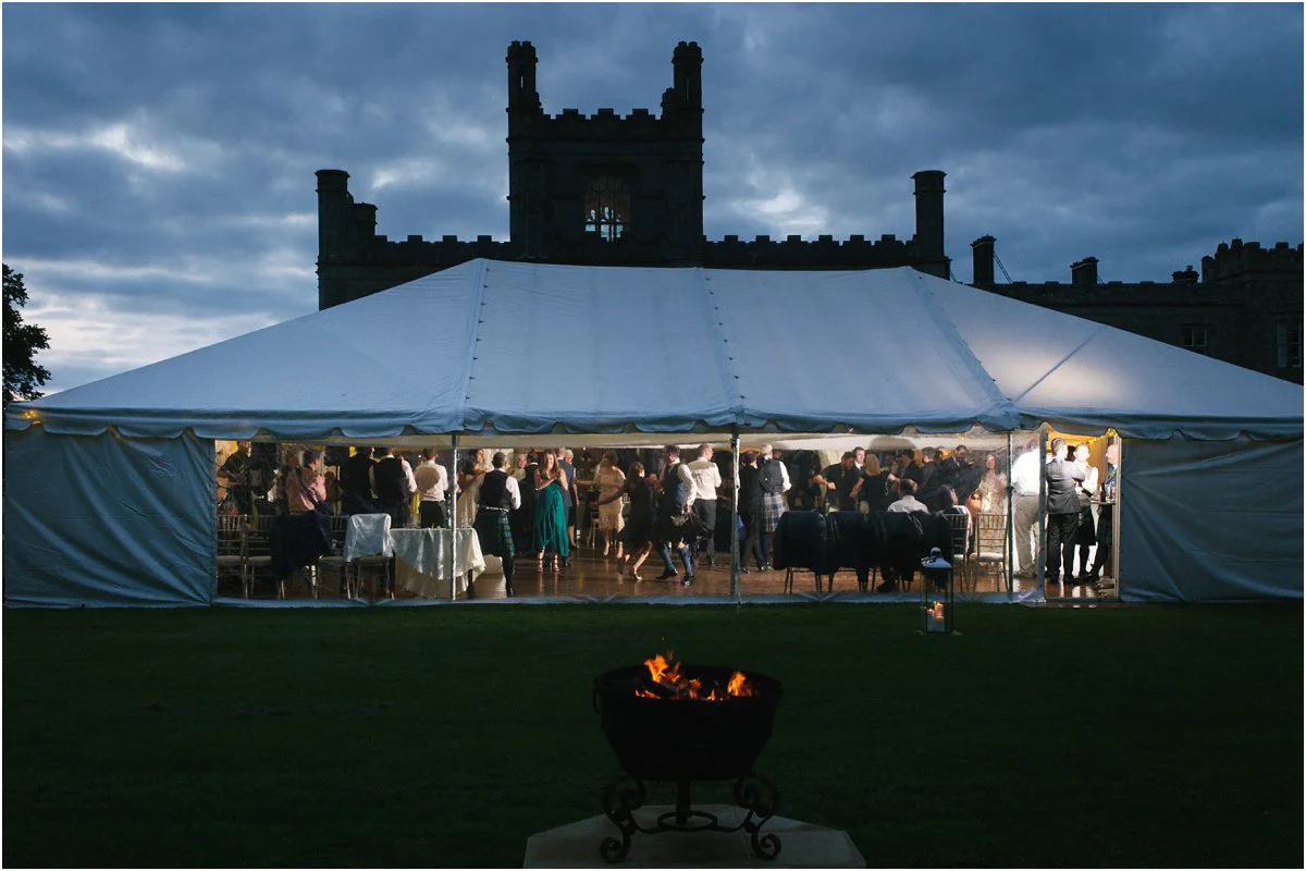 A lit marquee with translucent windows at dusk with a Scottish castle of Blairquhan during a summer wedding ceilidh dancing by Cro & Kow