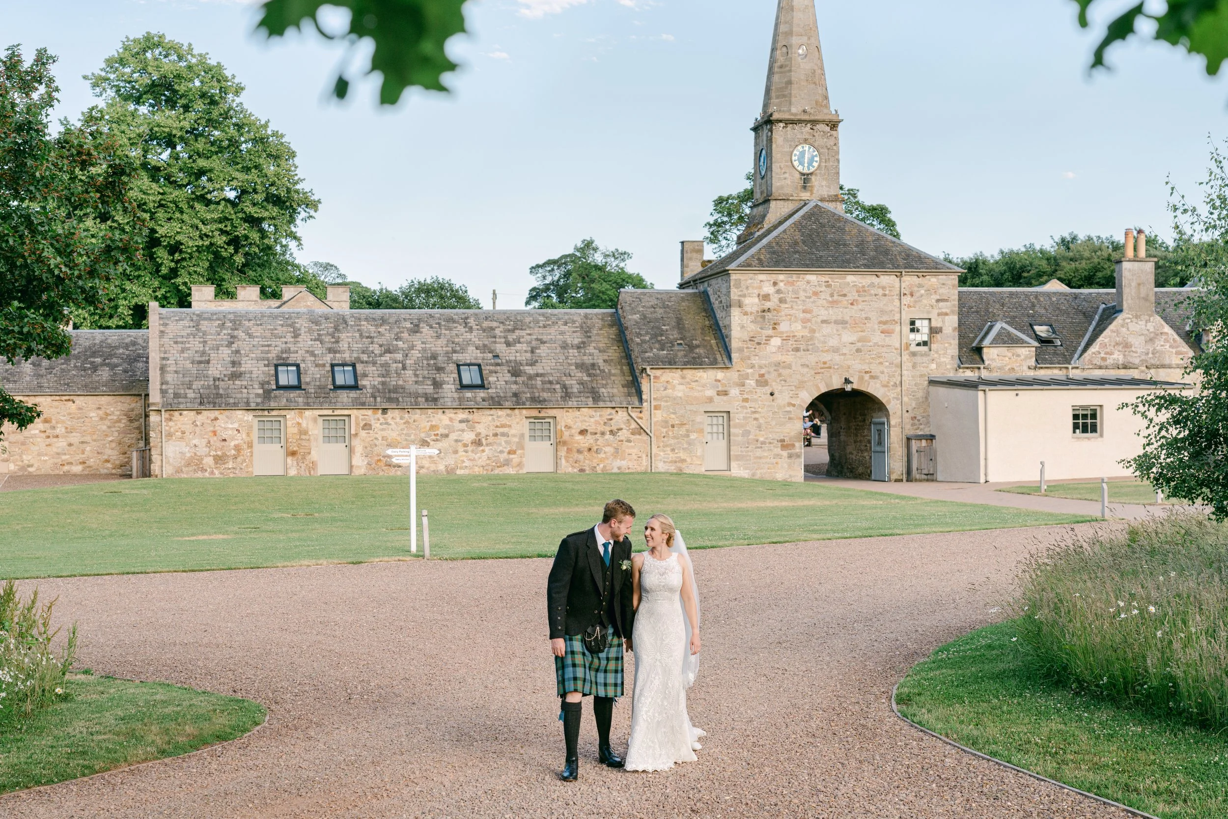 A couple walking in front of a historic stone building with a clock tower, surrounded by greenery. The man is wearing a kilt, and the woman is in a white wedding dress, indicating a wedding scene.