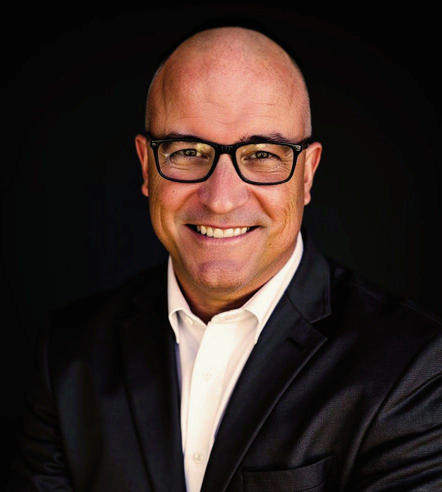 A smiling middle-aged man wearing glasses, a white collared shirt, and a black blazer, posed against a black background.