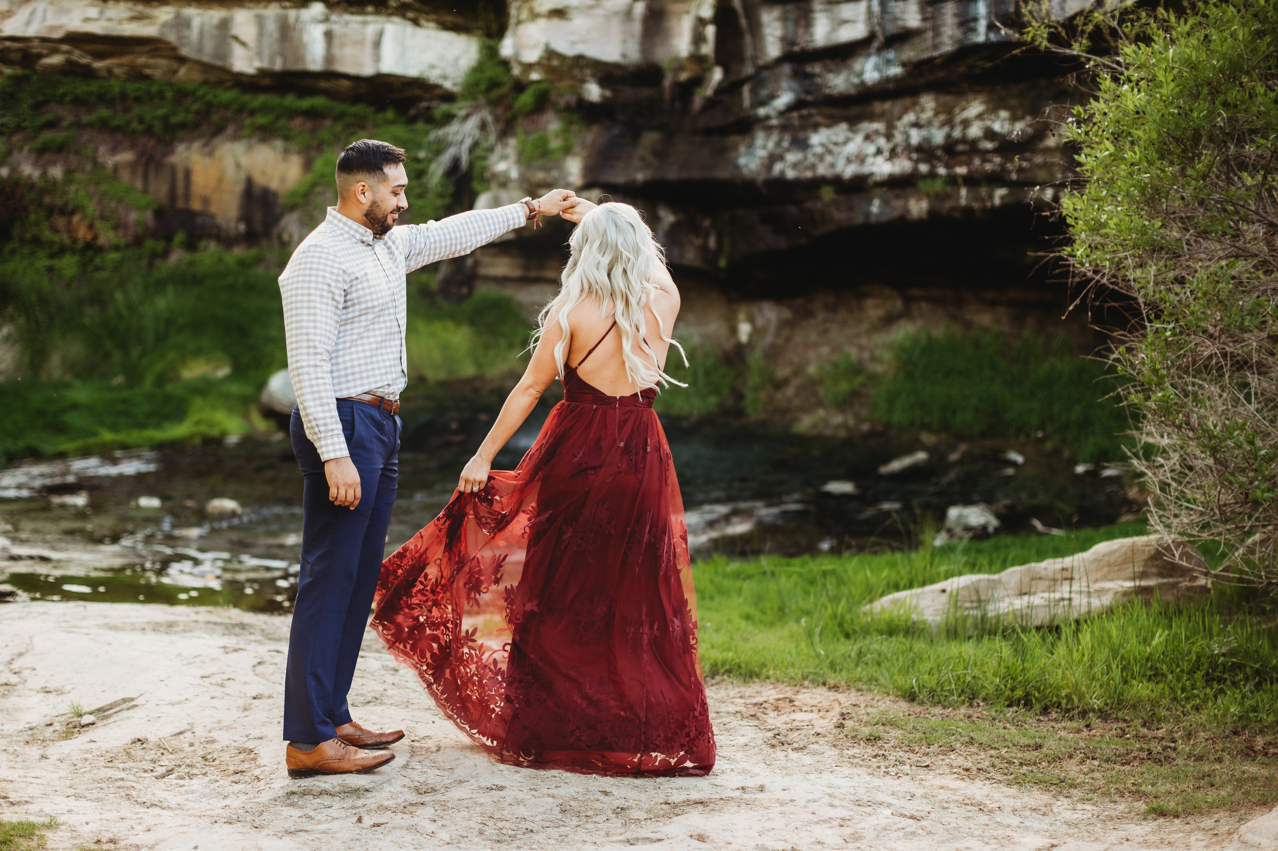 Engaged couple dancing in front of green foliage she is wearing a ruby red formal dress #engagementphotos #riverfalls #engaged #personality #amarillotexas #engagementphotographer #lifestylephotos #amarillophotographer #locationchoice #texasengagementphotos #engagment #tealawardphotography #wildliferefuge