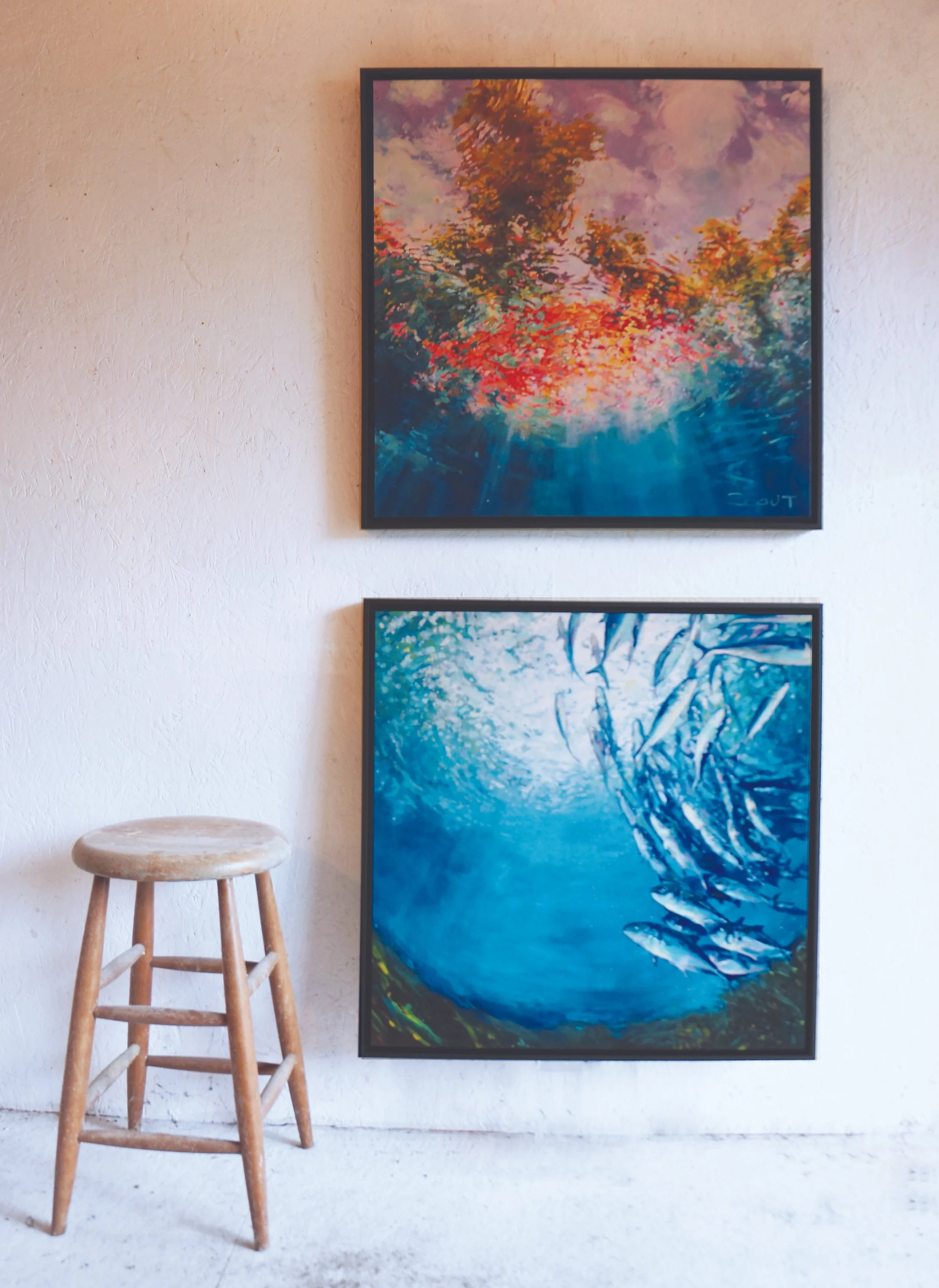 Two colorful abstract paintings on a beige wall, one above the other, with a wooden stool to the left.