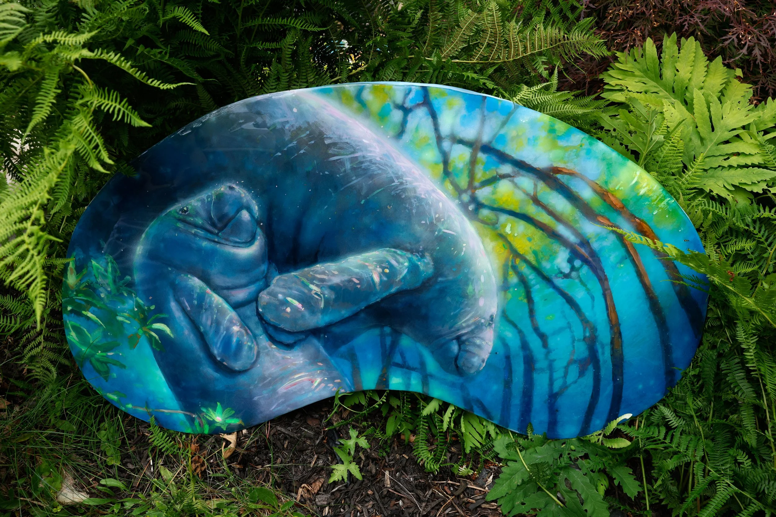 A painted rock with a mural of a hand and forest scene, surrounded by green plants and ferns.