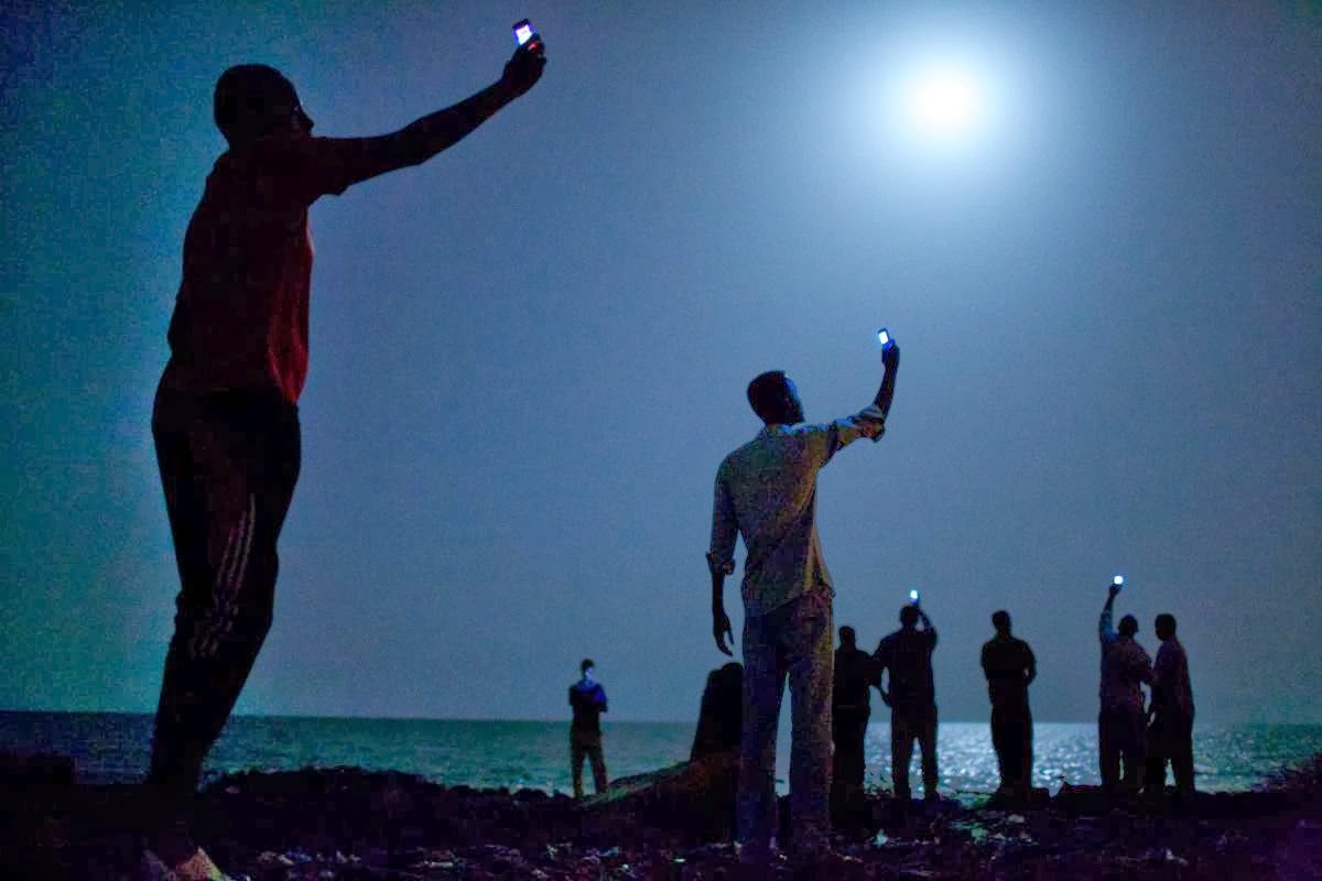 African migrants on the shore of Djibouti City at night raise their phones in an attempt to catch an inexpensive signal from neighbouring Somalia — a tenuous link to relatives abroad. (Credit: Jon Stanmeyer / VII).