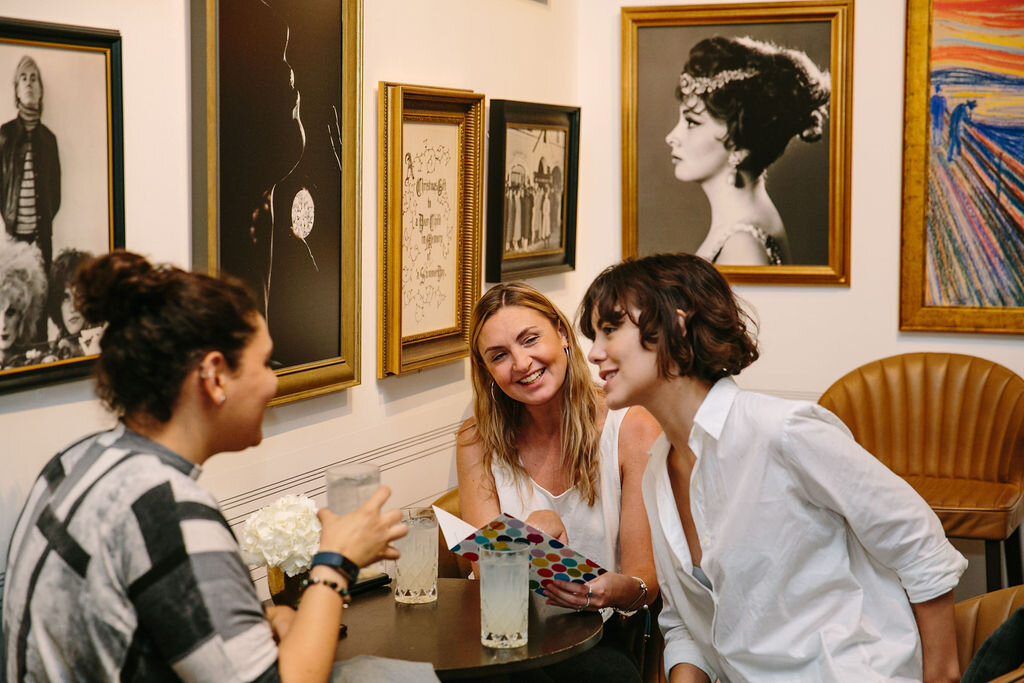 three pretty girls in cafe at gallery with art on walls and drinks in front of them