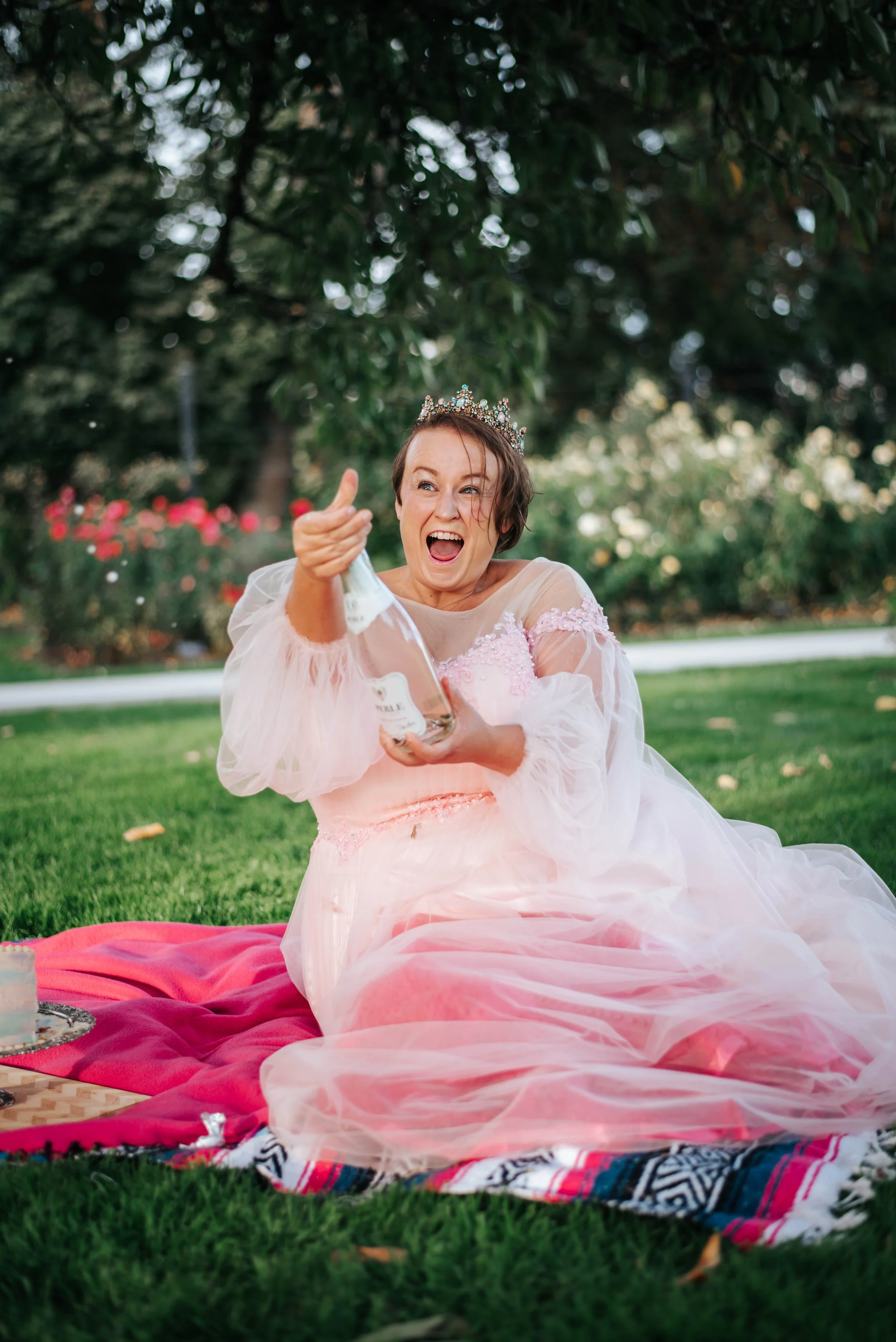 woman in a pink ball gown sitting in the park opening a bottle of sparkling wine