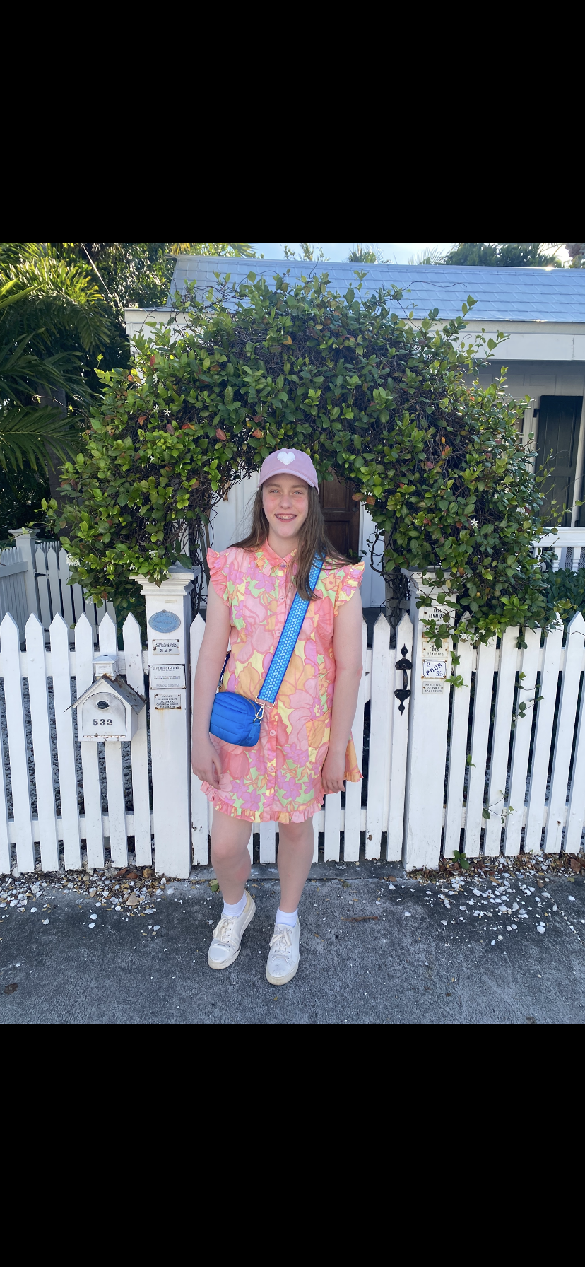 Image of white girl in floral dress and white tennis shoes in front of a green leafy archway and white picket fence