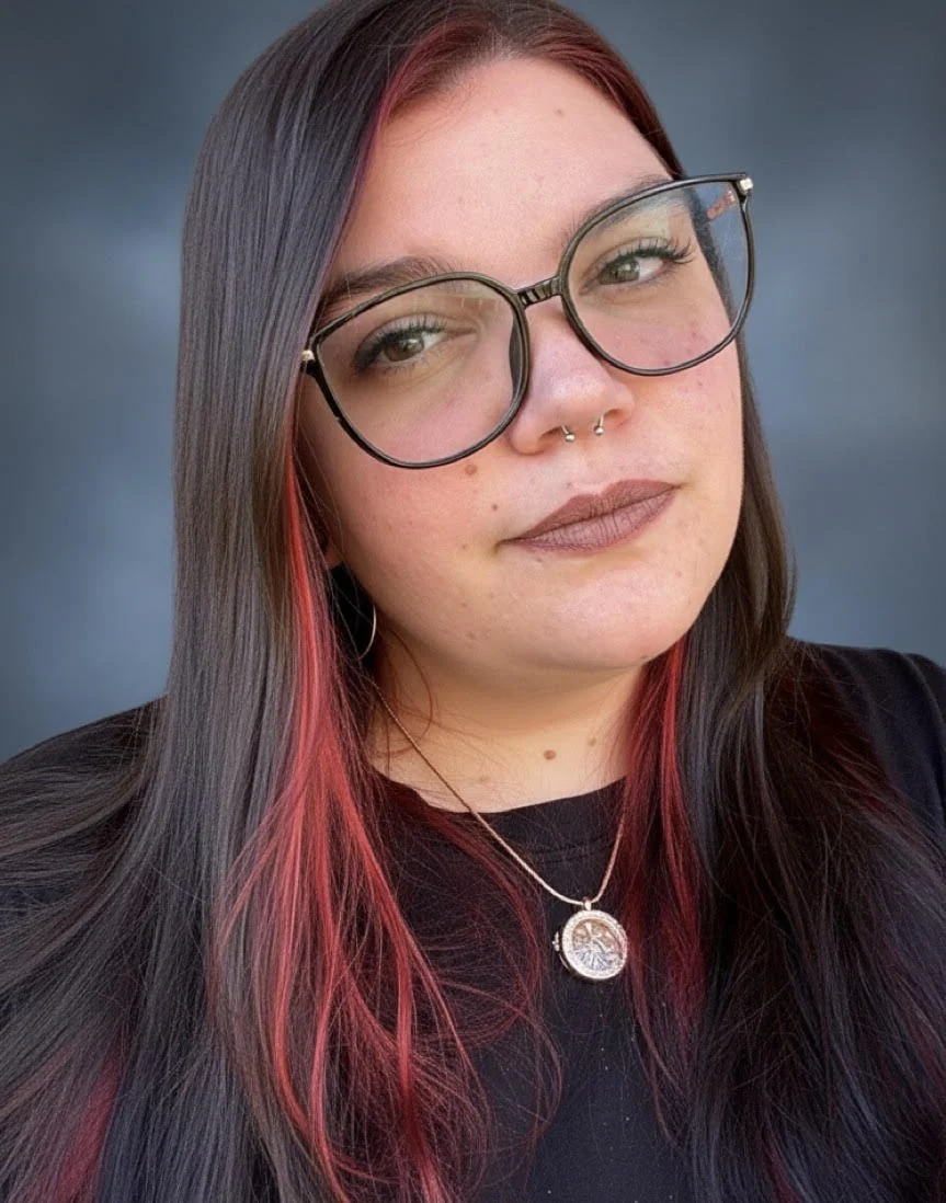 Light skinned woman with black frame glasses, septum piercing, and red streaks in her auburn hair