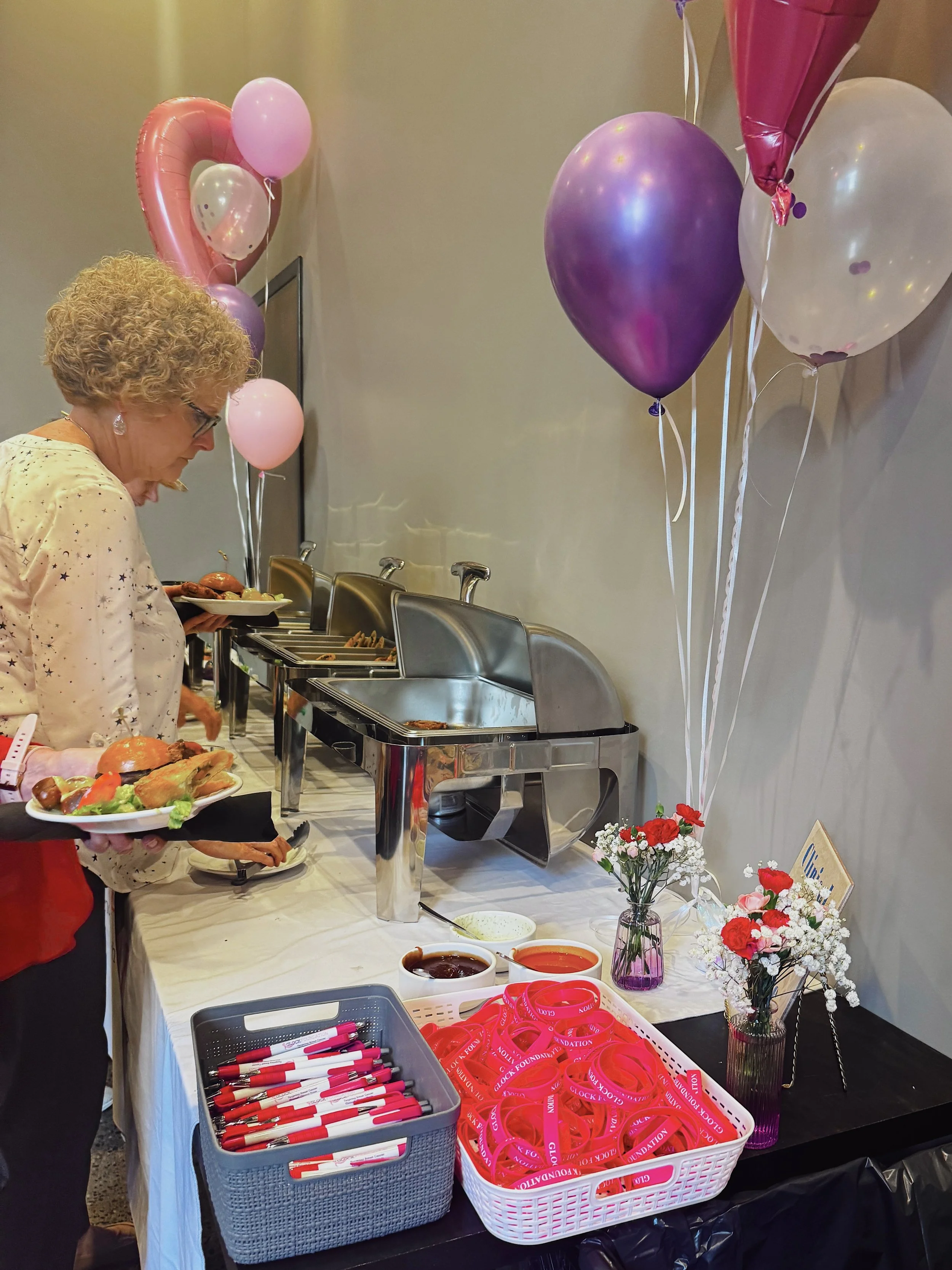 A woman at a buffet table with pink, purple, and white balloons, flowers, and red wristbands for a breast cancer awareness event.