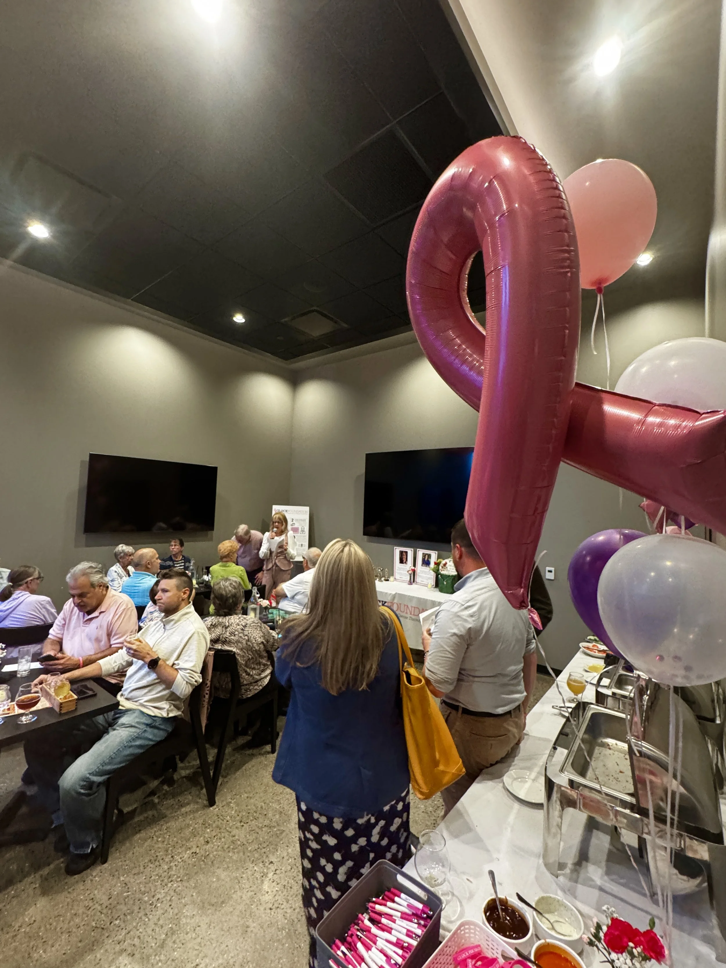 People gathered at a celebration with pink and purple balloons, a woman speaking at the front, and food on the buffet table.