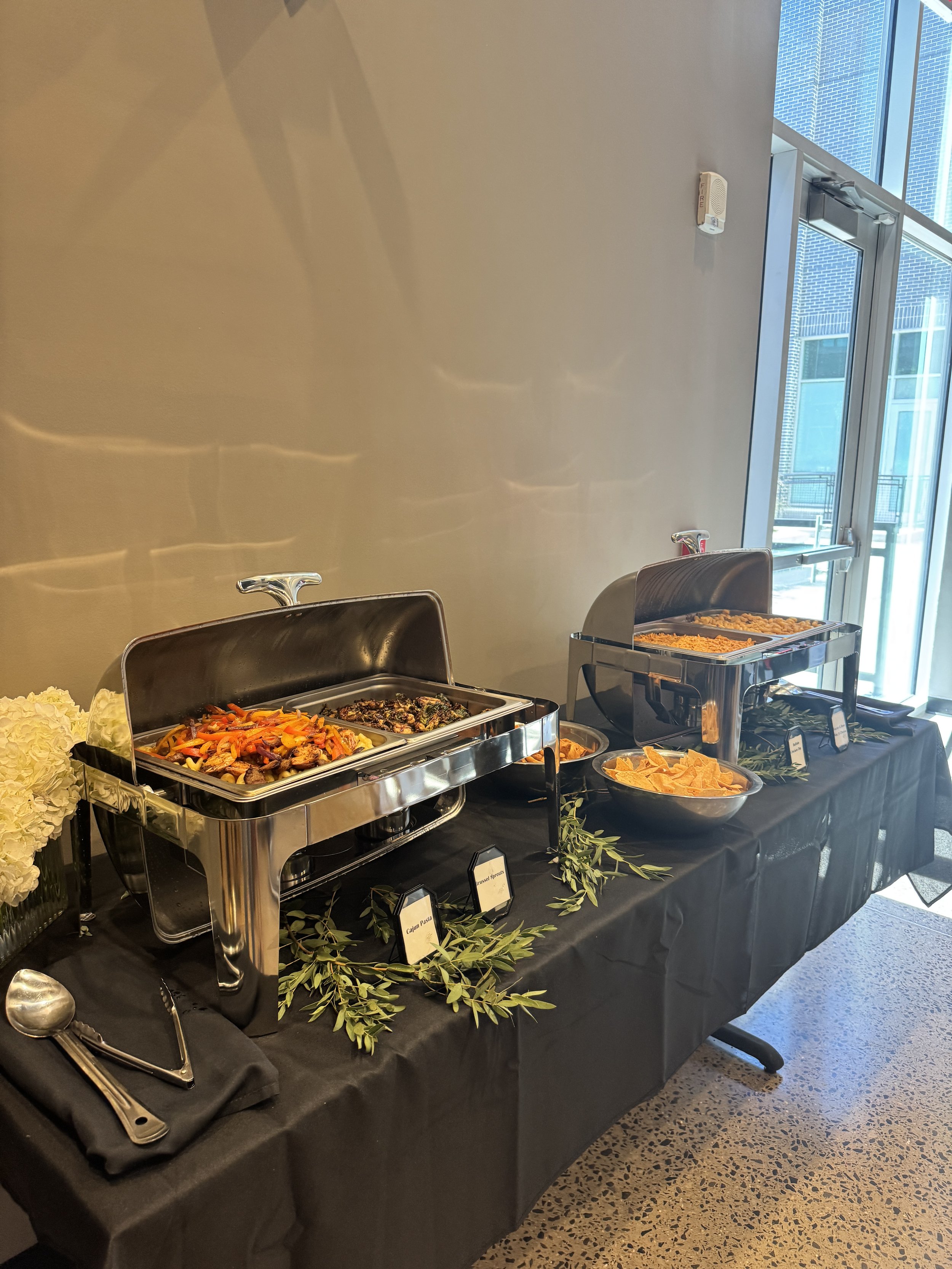Buffet table with covered chafing dishes containing dishes, bowls of chips, and labels on small signs. Black tablecloth, decorative greenery, and utensils are arranged on the table. Bright indoor space with large windows.