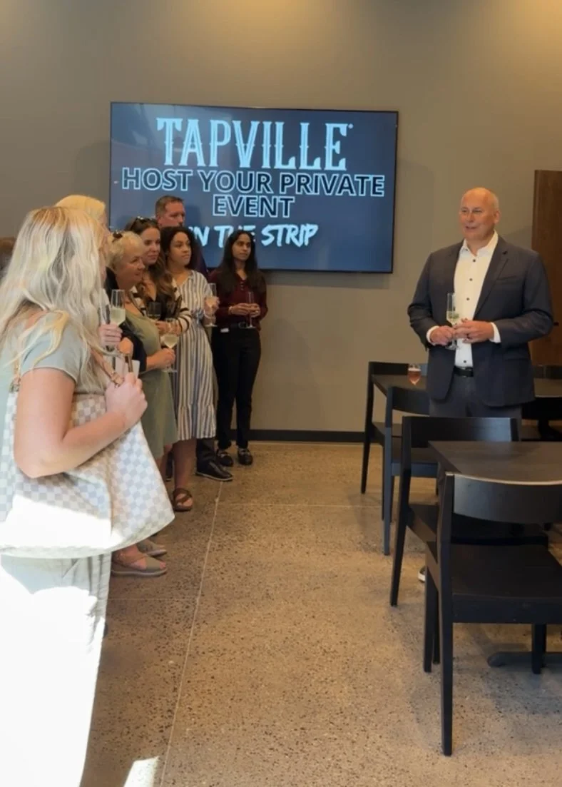 A group of people at a corporate event listening to a man in a suit giving a speech. Some hold champagne glasses. There is a large screen behind displaying an advertisement for Tapville.
