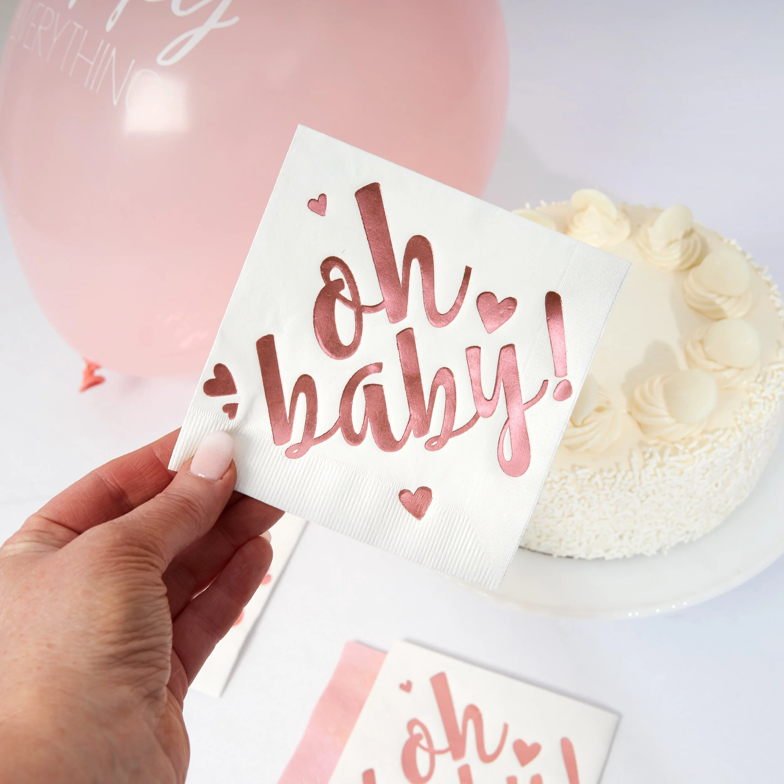 A hand holding a napkin with the words 'oh baby!' written in pink and red, surrounded by small heart illustrations. In the background, there is a white frosted cake decorated with white rosettes and a pink balloon.