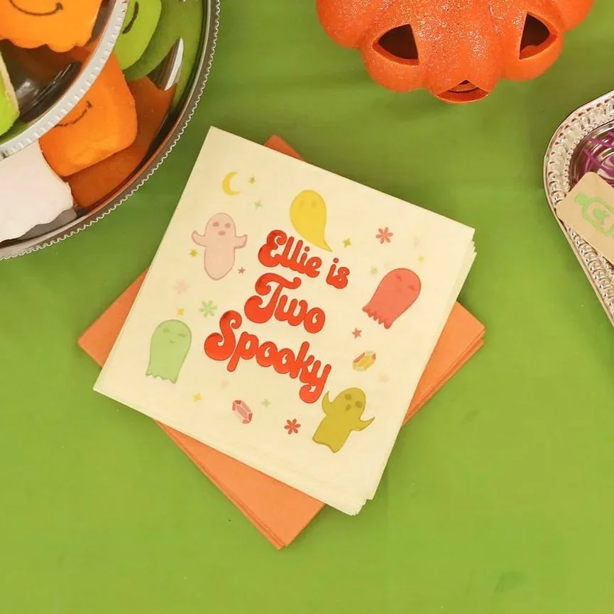 A paper napkin with colorful Halloween ghosts and the text "Ellie is Two Spooky" on a green table.